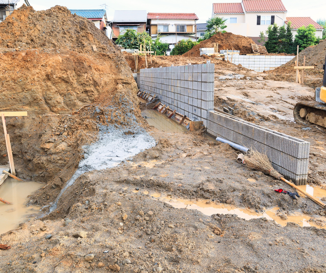 Construction site with a retaining wall made of concrete blocks. Muddy ground surrounds the wall, with dirt piles in the background.