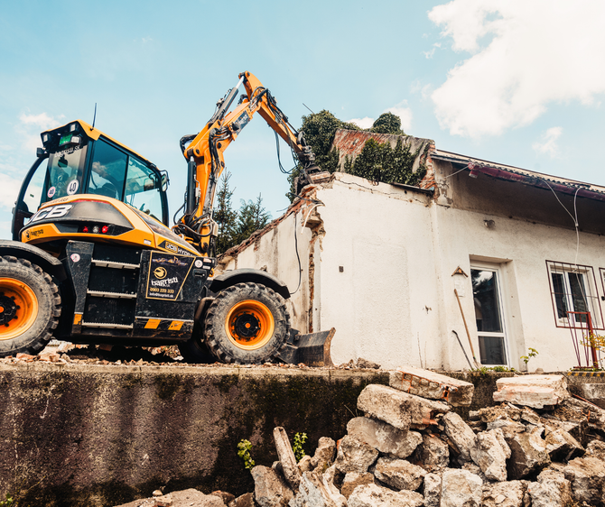 Yellow excavator demolishing a white building under a cloudy sky.