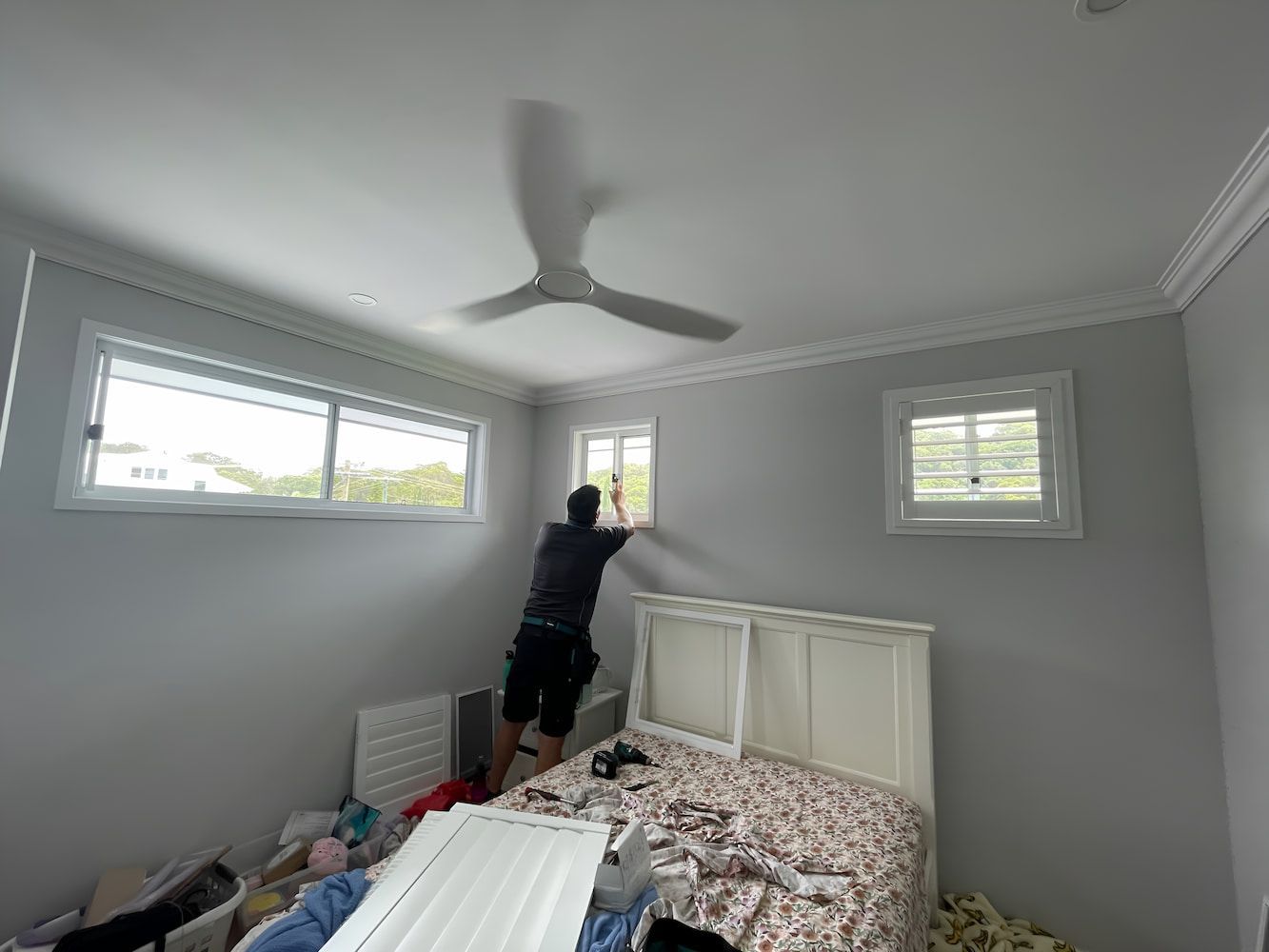 A Man Is Cleaning A Window In A Bedroom With A Ceiling Fan — Timeless Blinds And Shutters In Burleigh Heads, QLD