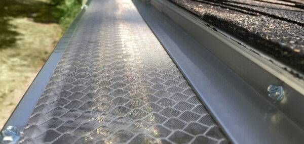 Close-up of a gutter with a mesh screen. The gutter is attached to a roof with asphalt shingles.