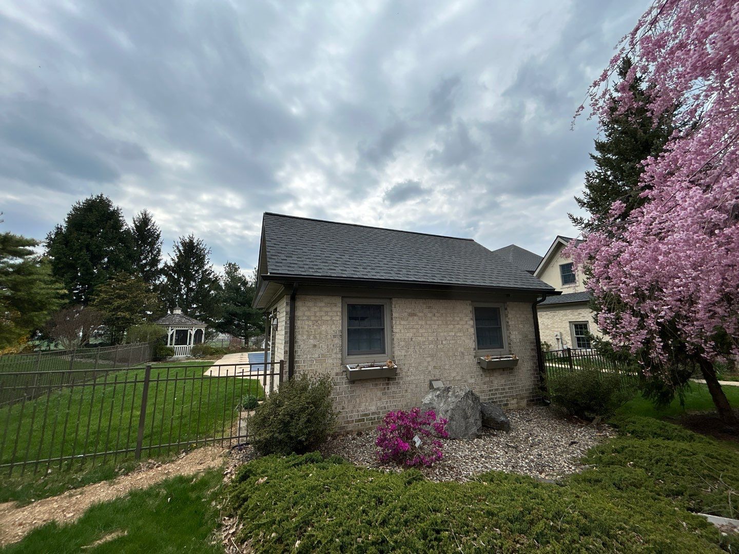 Small stone building with a dark roof and window boxes, next to a pink flowering tree and green lawn under a cloudy sky.