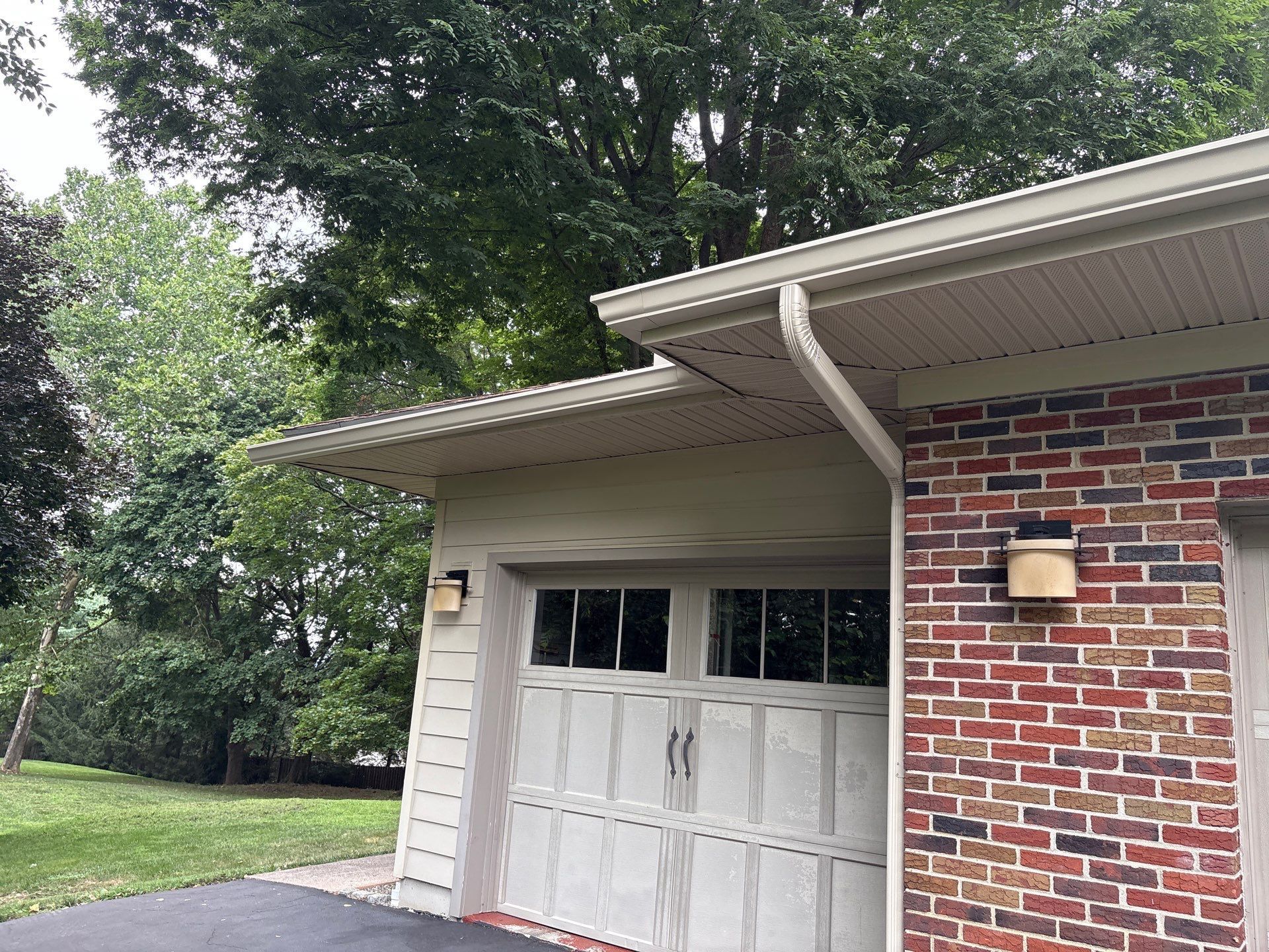 Two-story house with tan siding, black shutters, white porch with hanging plant, and a cloudy sky.