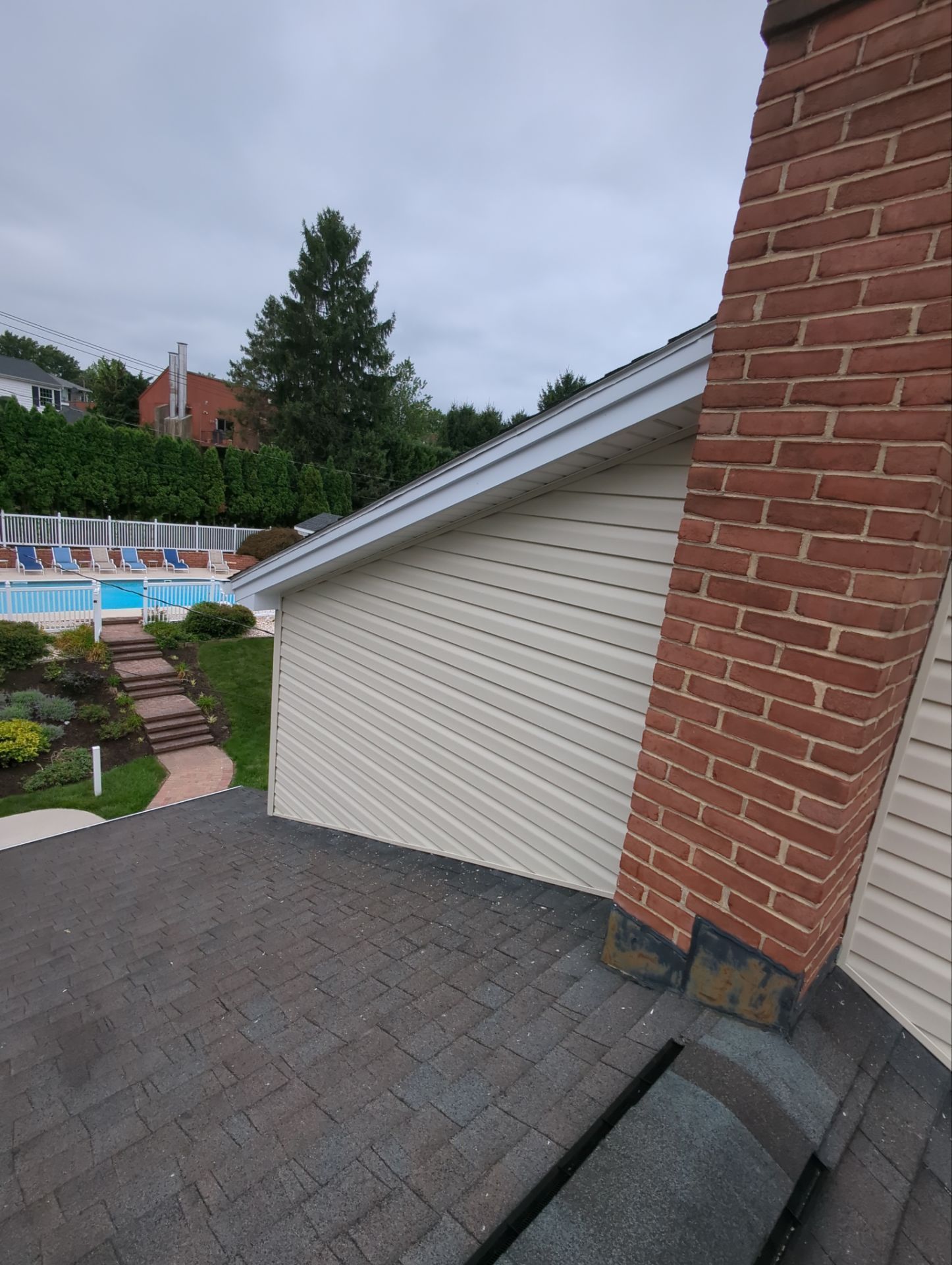 Tan siding next to a brick chimney and dark roof. A pool area and trees are visible in the background.