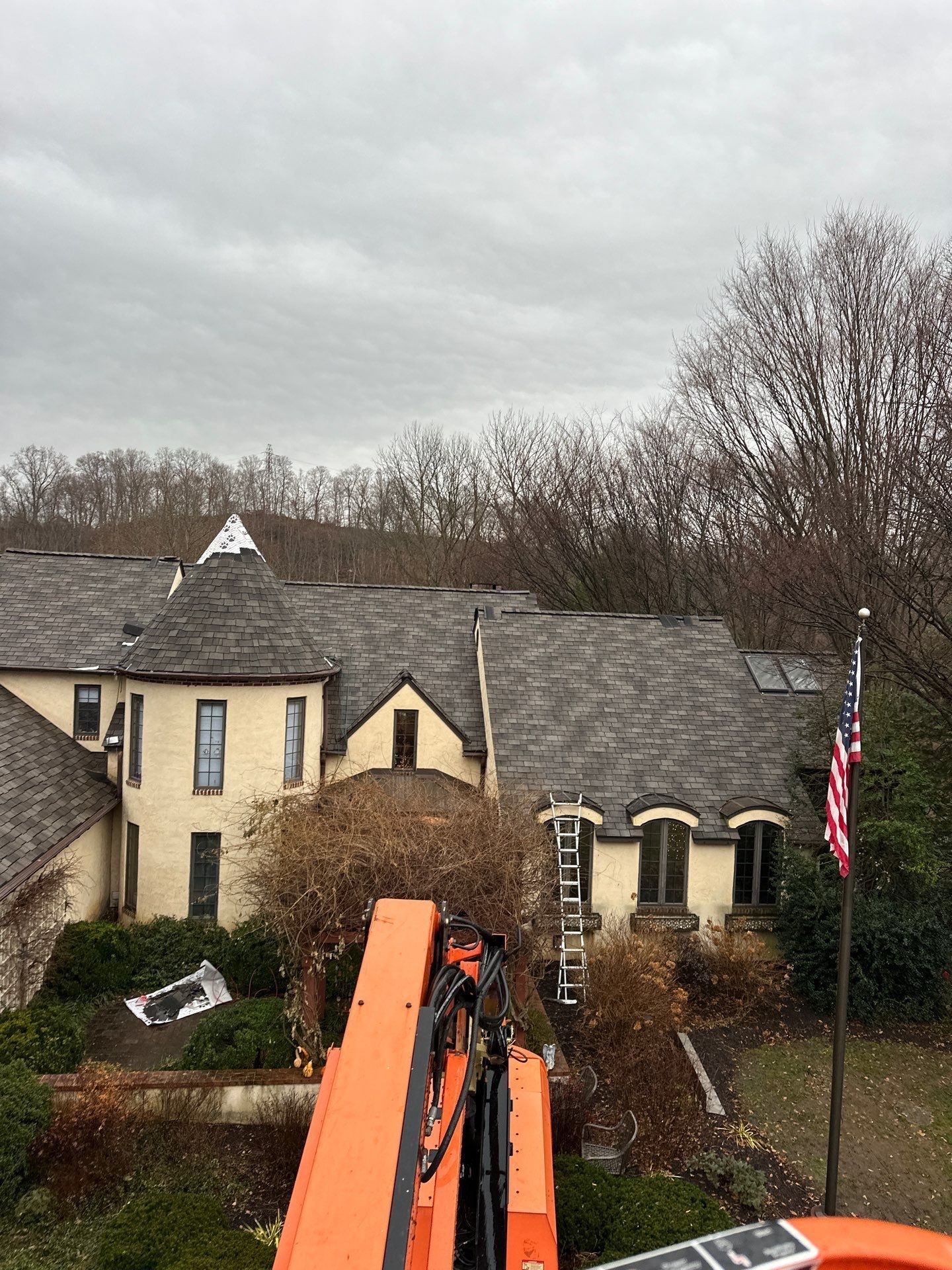 House with a gray roof and beige walls, viewed from a crane. An American flag flies to the right. Overcast sky.