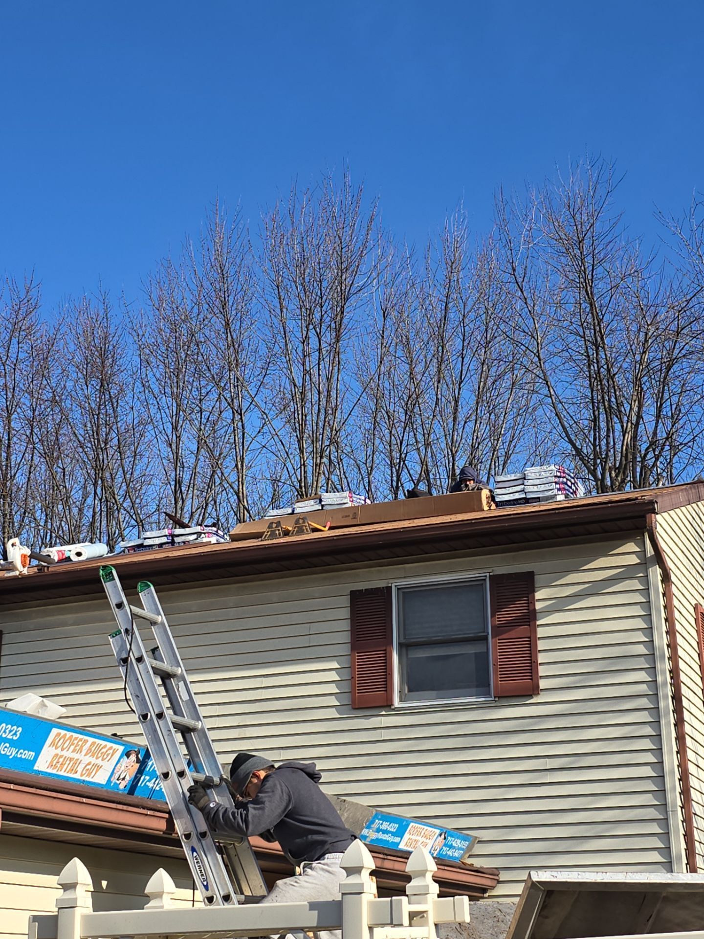 Roofer on a ladder with materials on a roof against a clear blue sky.