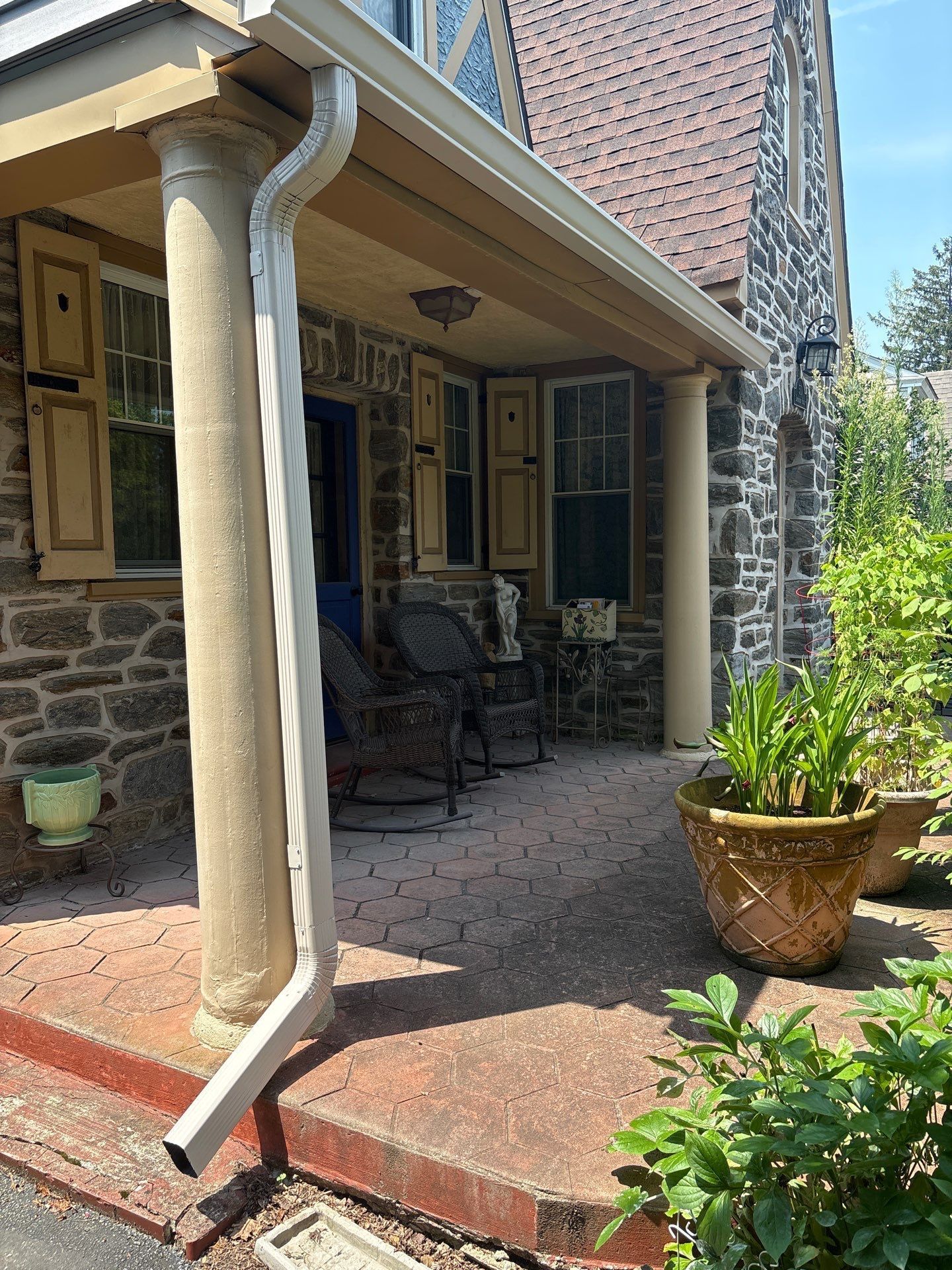 Stone house porch with columns, shutters, seating, and potted plants.