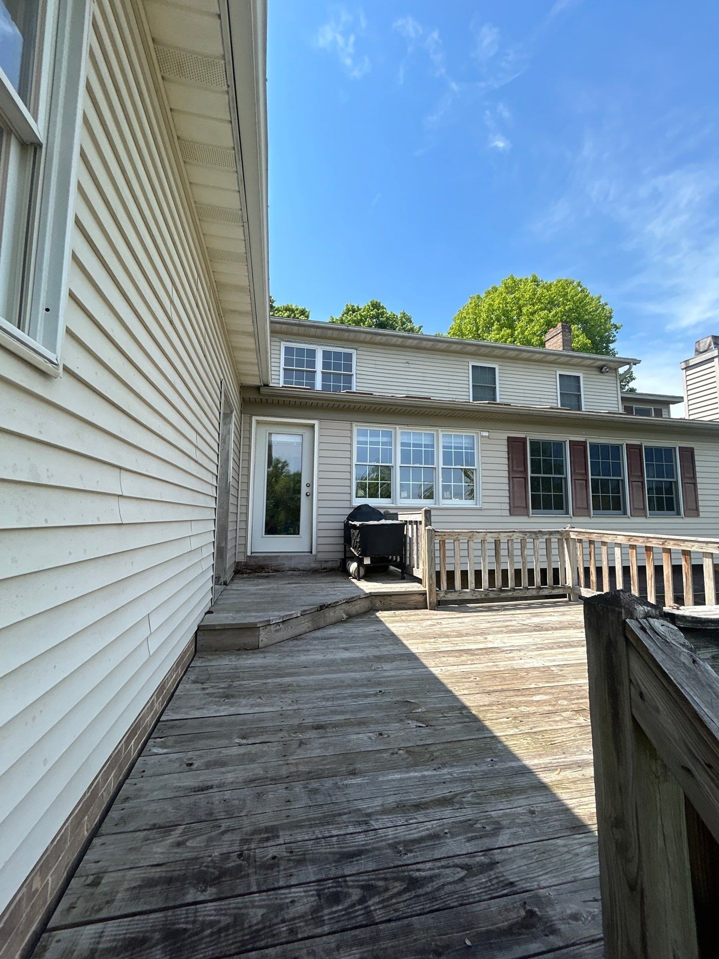 Wooden deck extends to door, grilling area, two-story house, sunny day, blue sky.