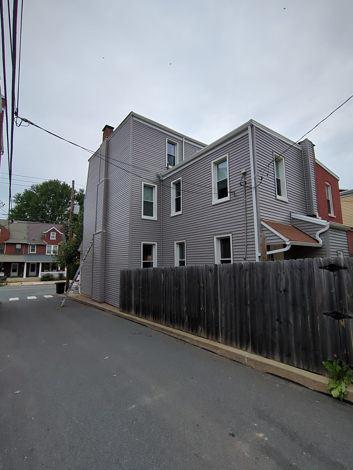 Two-story house with tan siding, black shutters, white porch with hanging plant, and a cloudy sky.