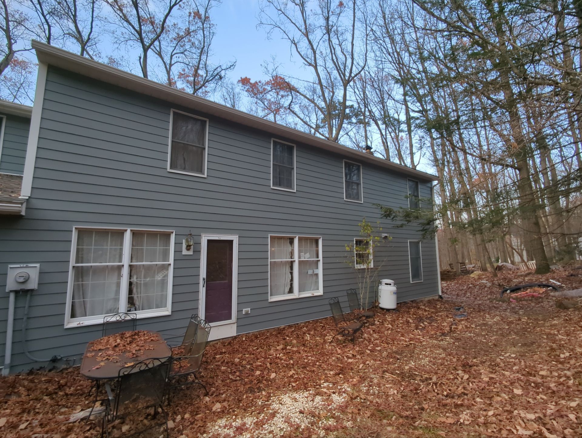 Two-story gray house with white window frames, surrounded by trees and fallen leaves.