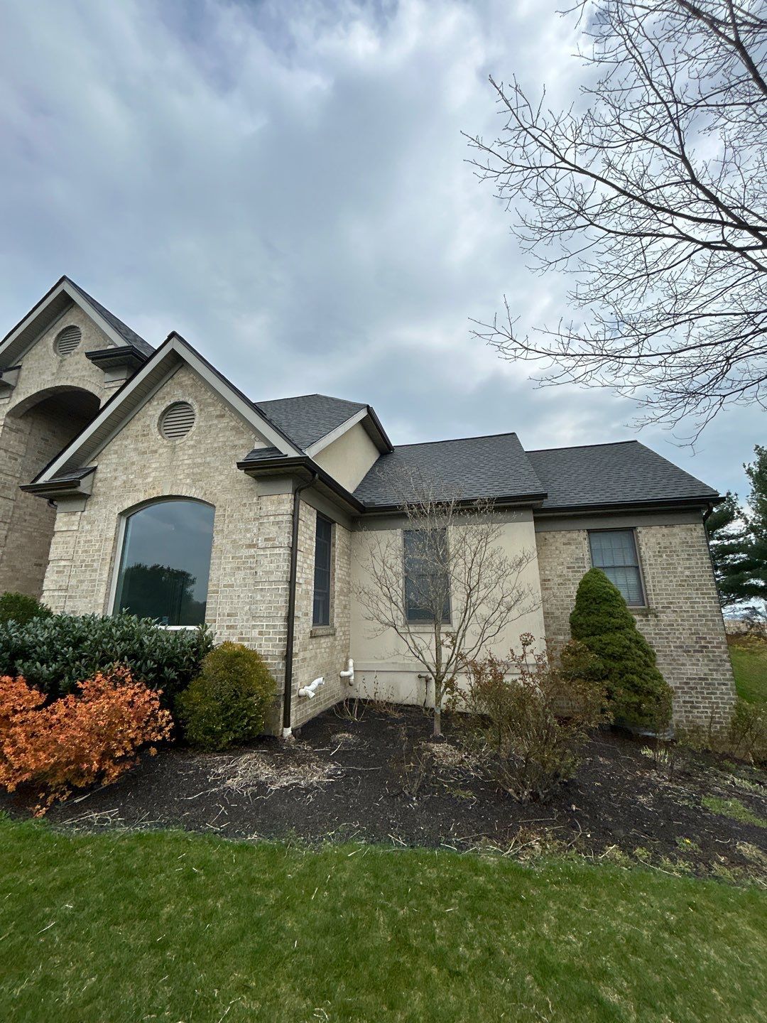 Beige brick house with dark gray roof against a cloudy sky. Landscaping in front.