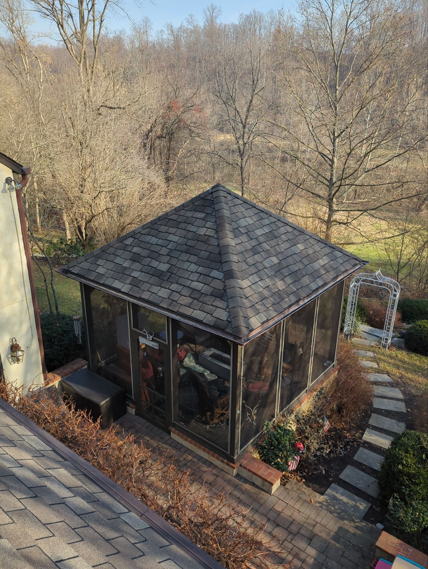 Screened-in gazebo with a dark shingled roof, set on a stone path with a backdrop of bare trees.