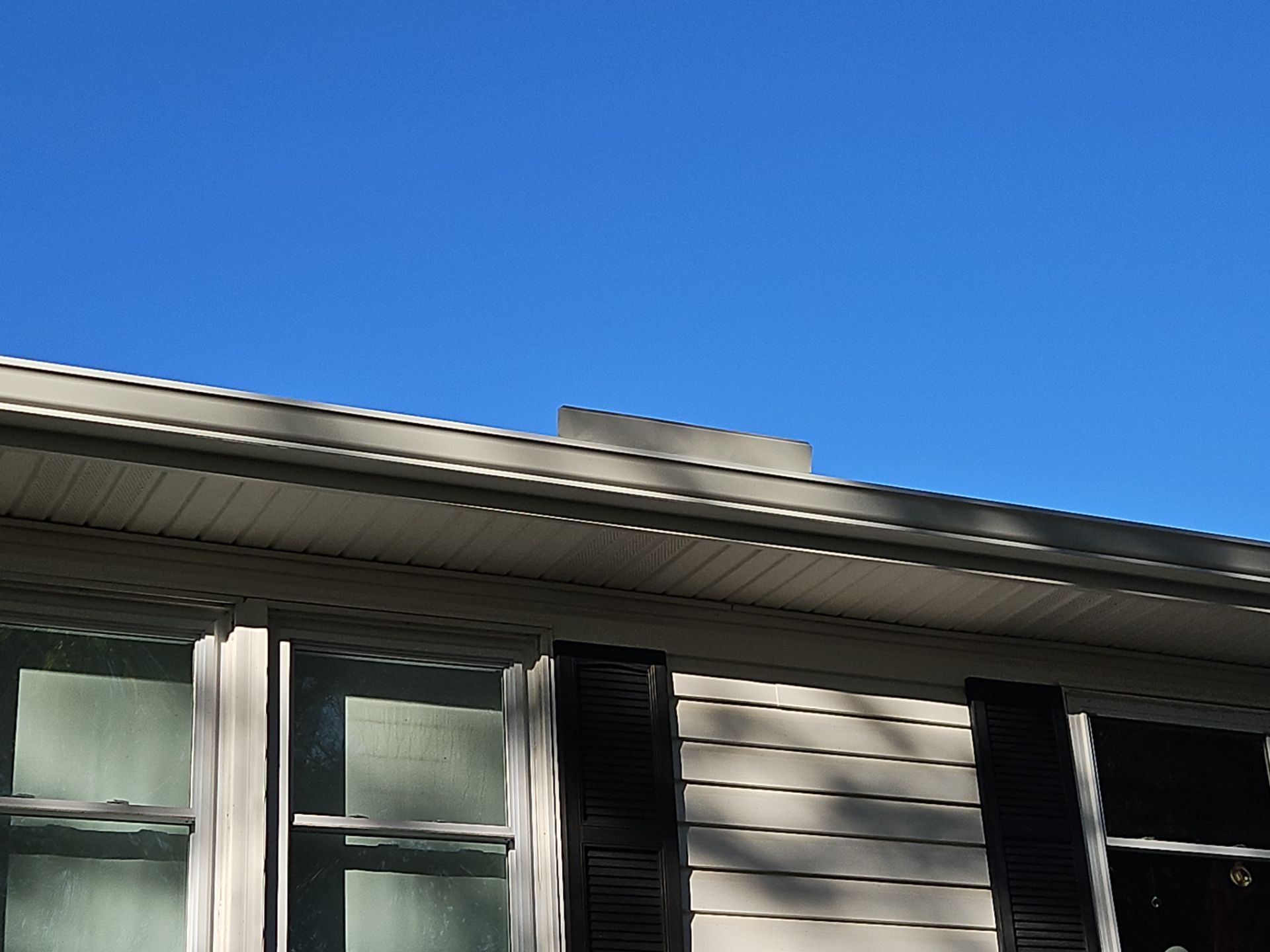 Close-up of a house exterior with light siding, black shutters, and a dark vent against a bright blue sky.