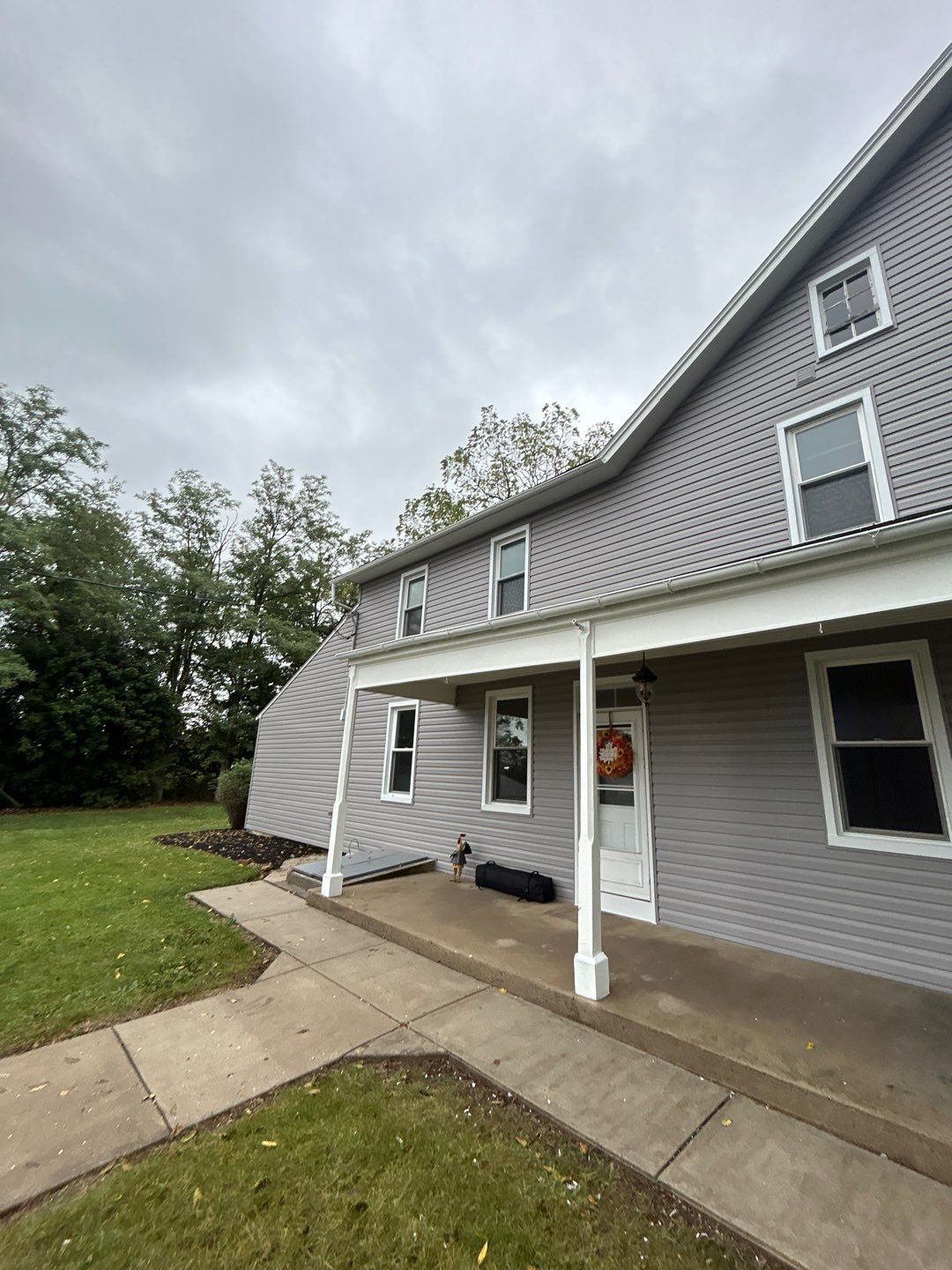 Two-story house with tan siding, black shutters, white porch with hanging plant, and a cloudy sky.