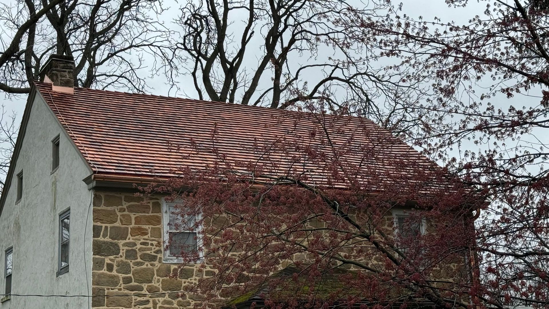 Stone house with wavy brown roof, surrounded by trees with pink blooms and bare branches against a cloudy sky.
