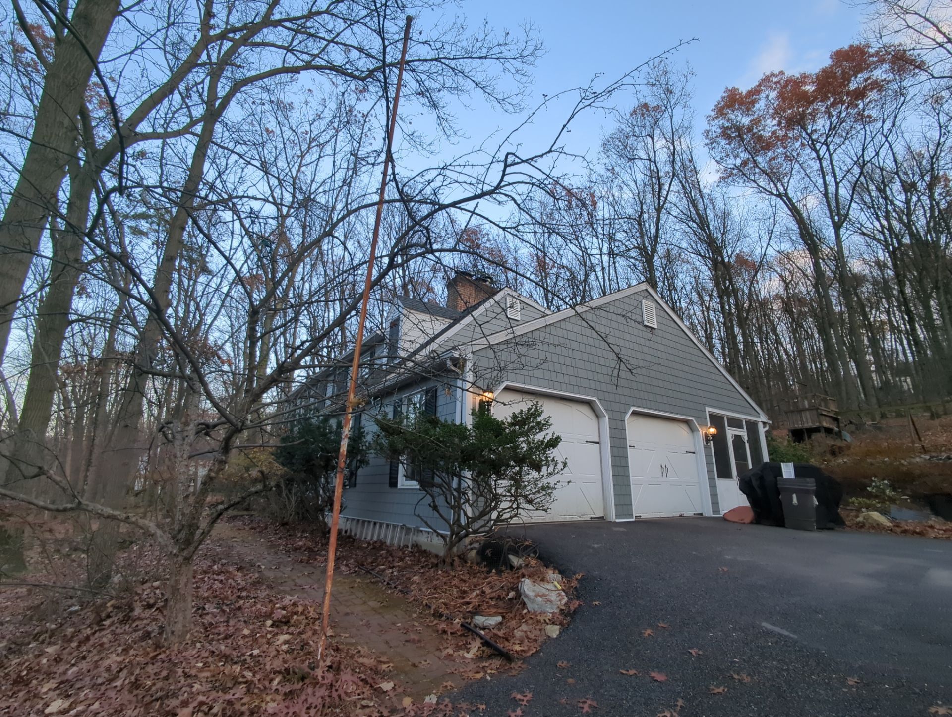 Gray house with a two-car garage, surrounded by bare trees, on a driveway leading uphill.