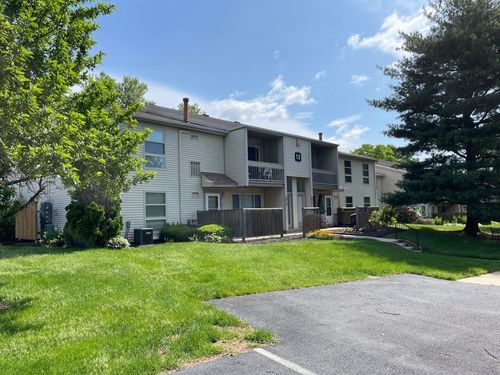 Two-story beige apartment building with balconies, green lawn, trees, and blue sky.
