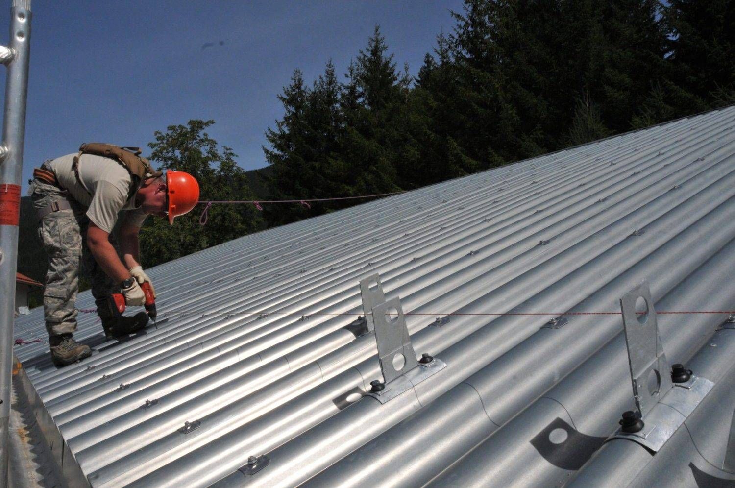 Person in an orange hard hat works on a corrugated metal roof, with safety equipment attached. Forest visible in background.