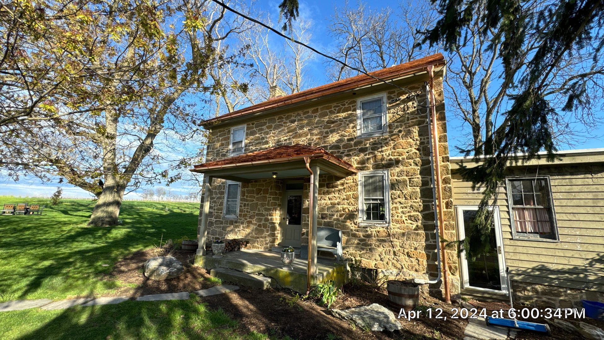 Two-story stone house with a covered porch and small windows. A green lawn and trees surround the building.