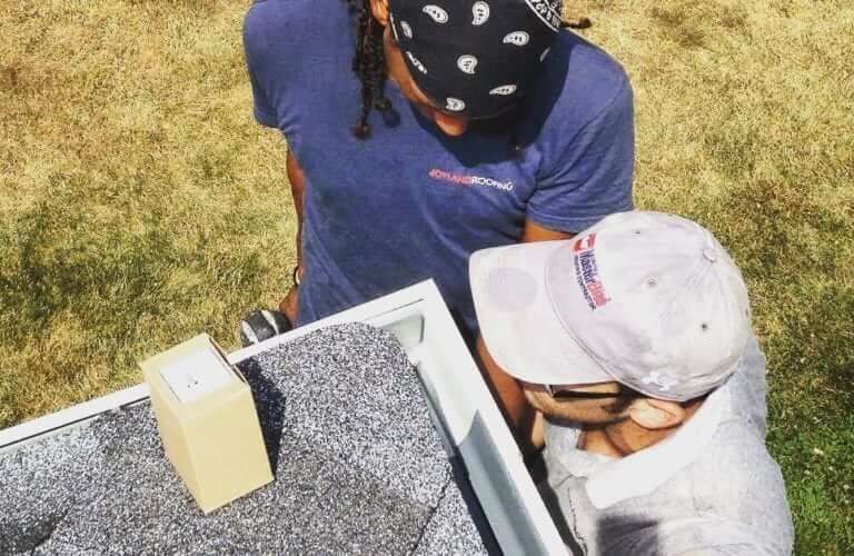 Two people work on a roof, one in a bandana, the other in a cap, near a box and a gutter.