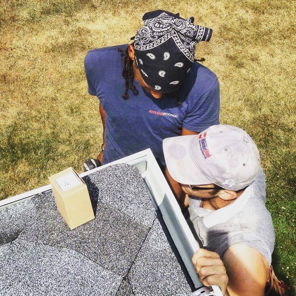 Two people installing a gutter on a roof outdoors; one wearing a bandana, the other a baseball cap.