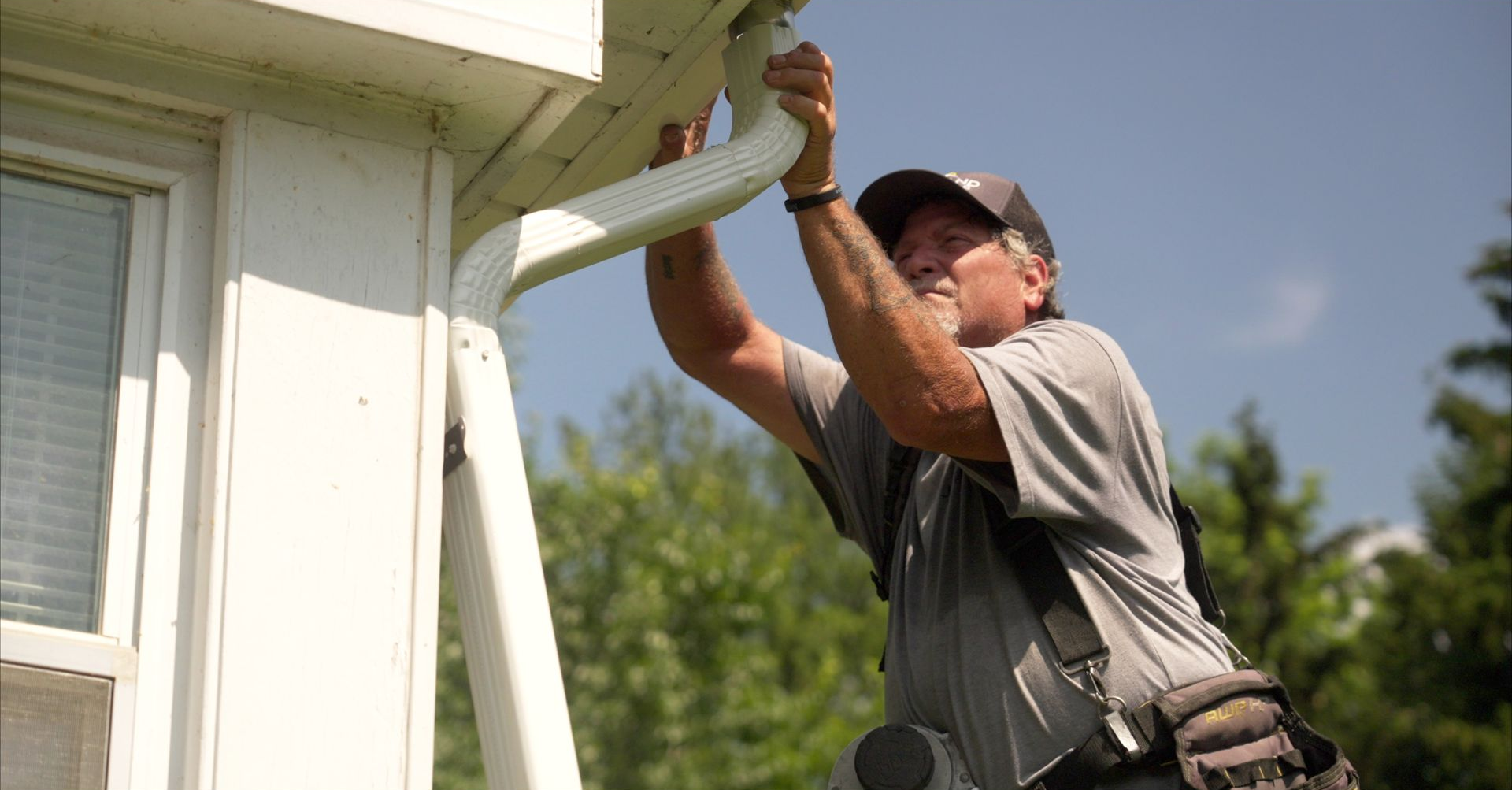 Man on ladder installing gutter on a white house under a blue sky.