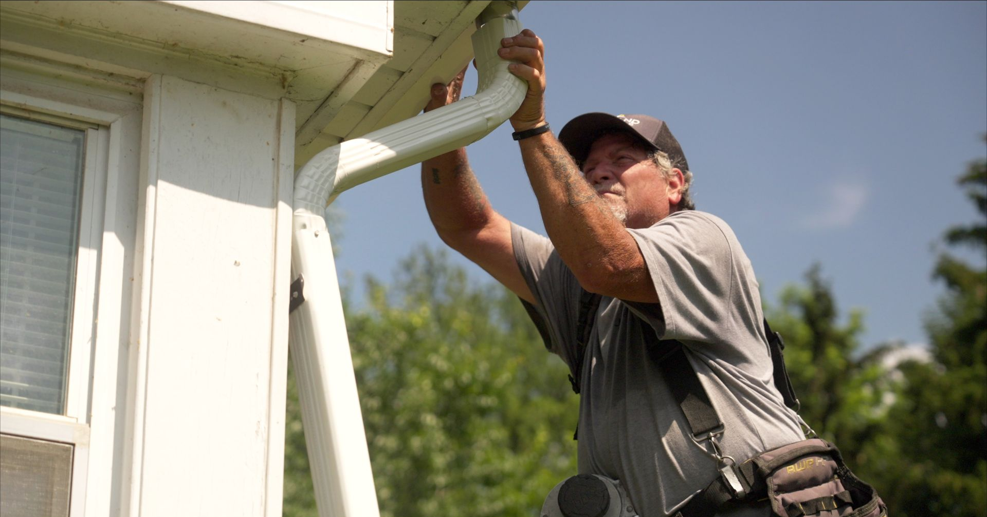 Man on a ladder installing a white gutter on a white house under a blue sky.