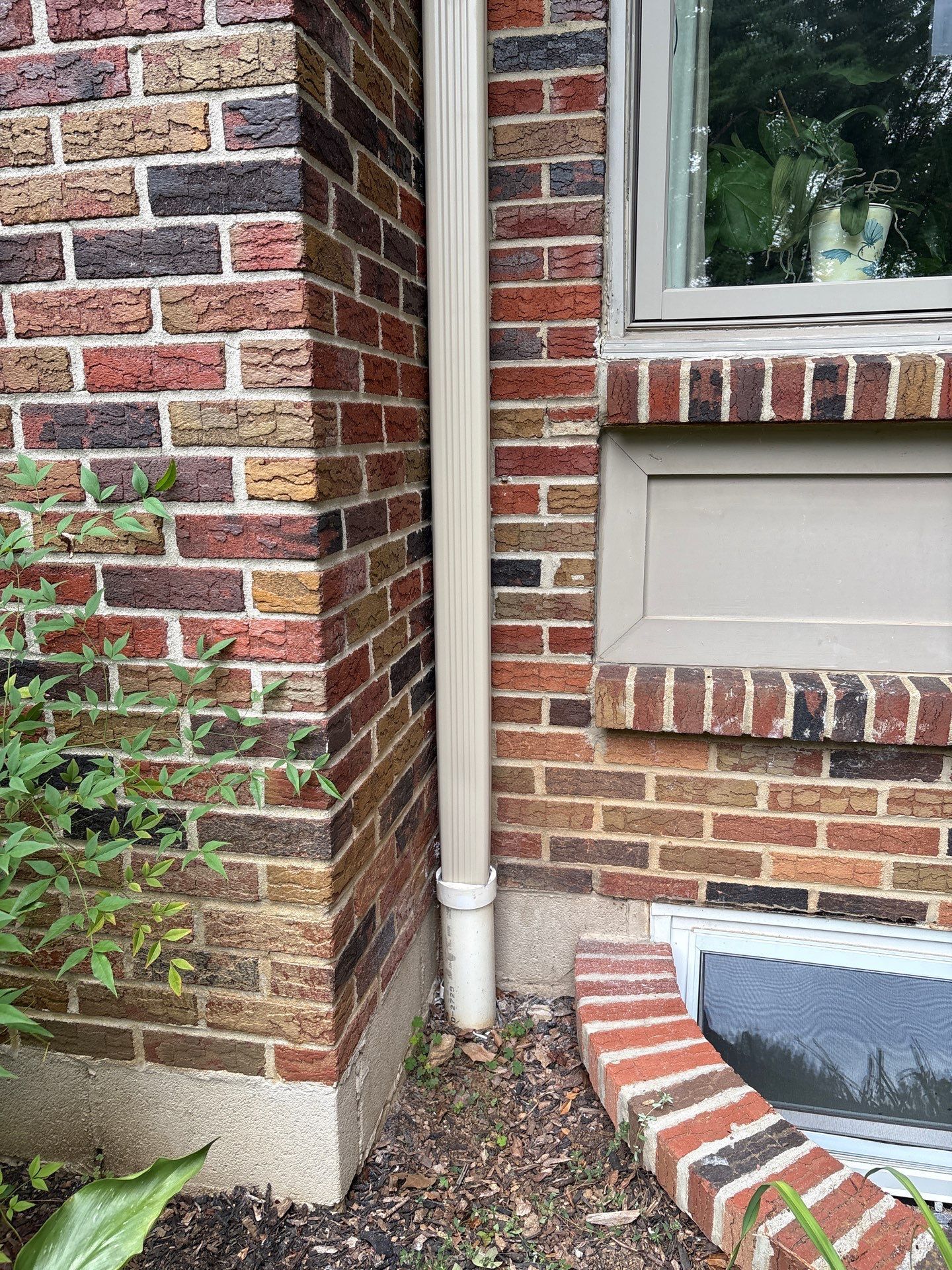 Brick building corner with a beige downspout and a small window with a brick window well.