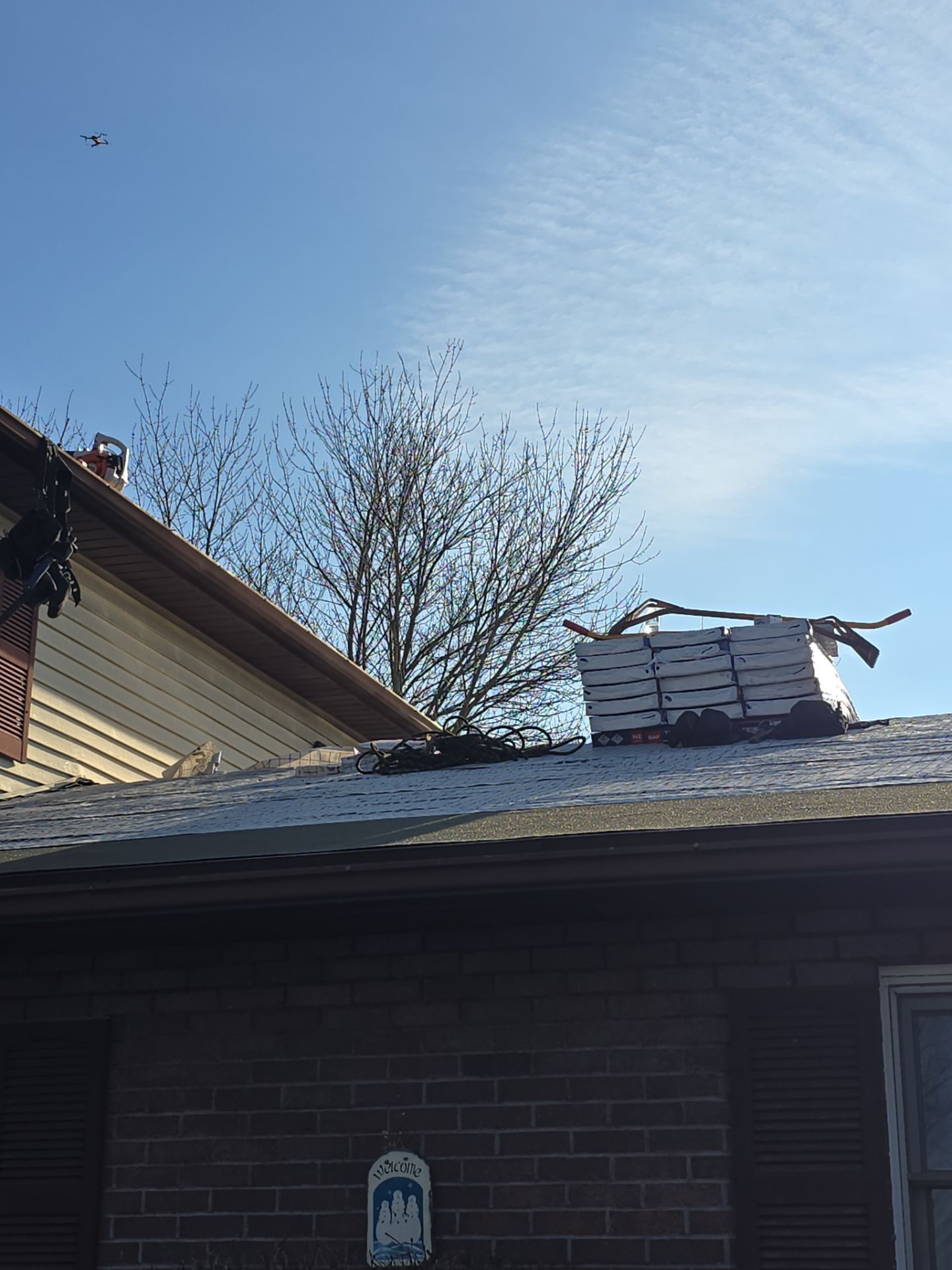Roof with stacked tiles, branches, and a brick house against a blue sky.