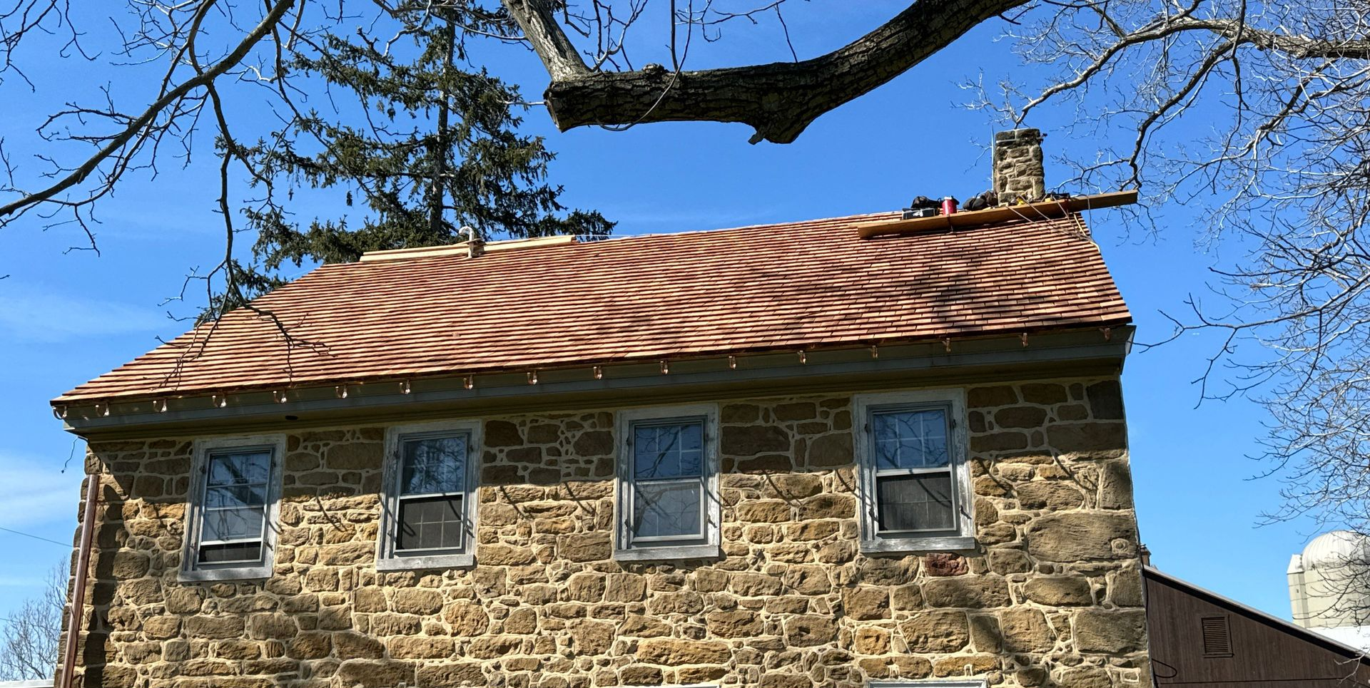 Stone house with wavy copper roof, windows, and chimney against a blue sky, branches overhead.