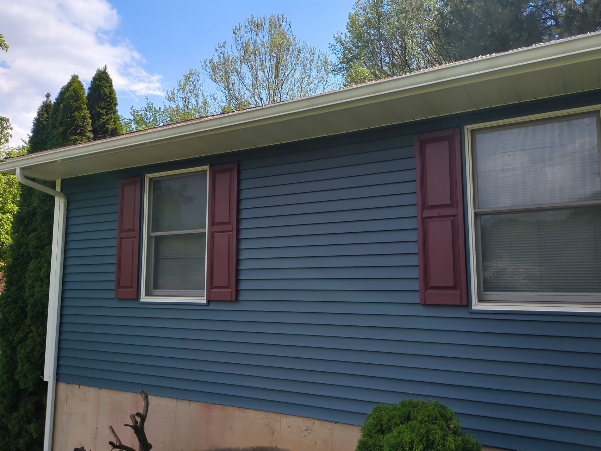 Two-story house with tan siding, black shutters, white porch with hanging plant, and a cloudy sky.
