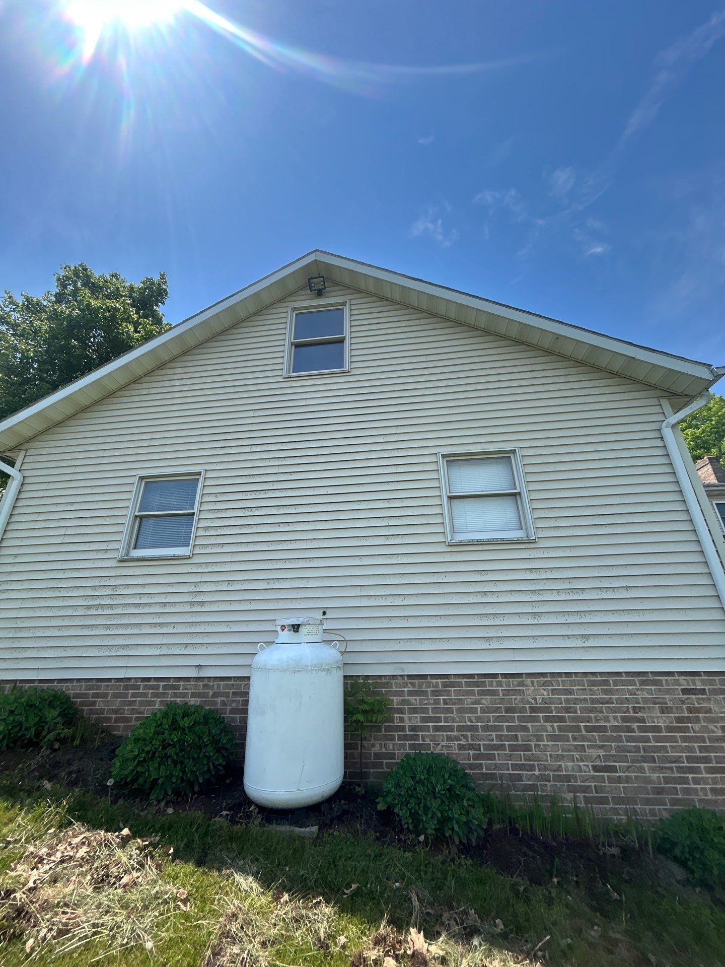 White-sided house with three windows, a propane tank, and brick foundation, under a bright blue sky.