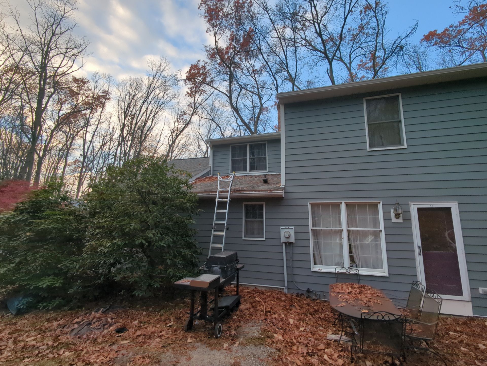 Gray house with a ladder propped against the roof. Fall leaves cover the ground.
