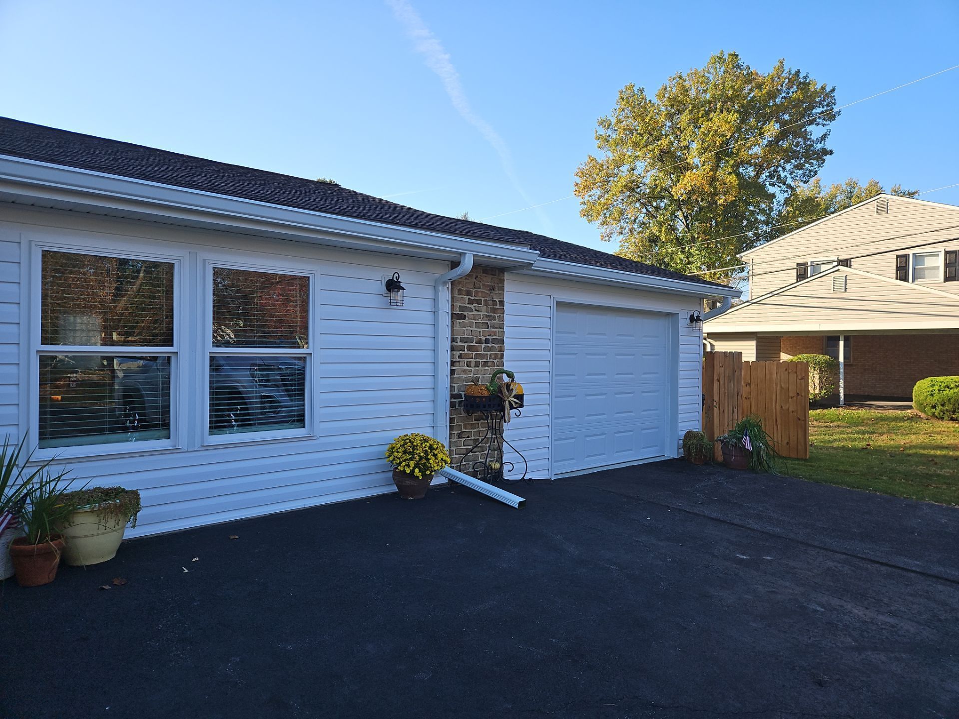 White house with garage and driveway. Brick accent, flower pots, and a fence. Blue sky.