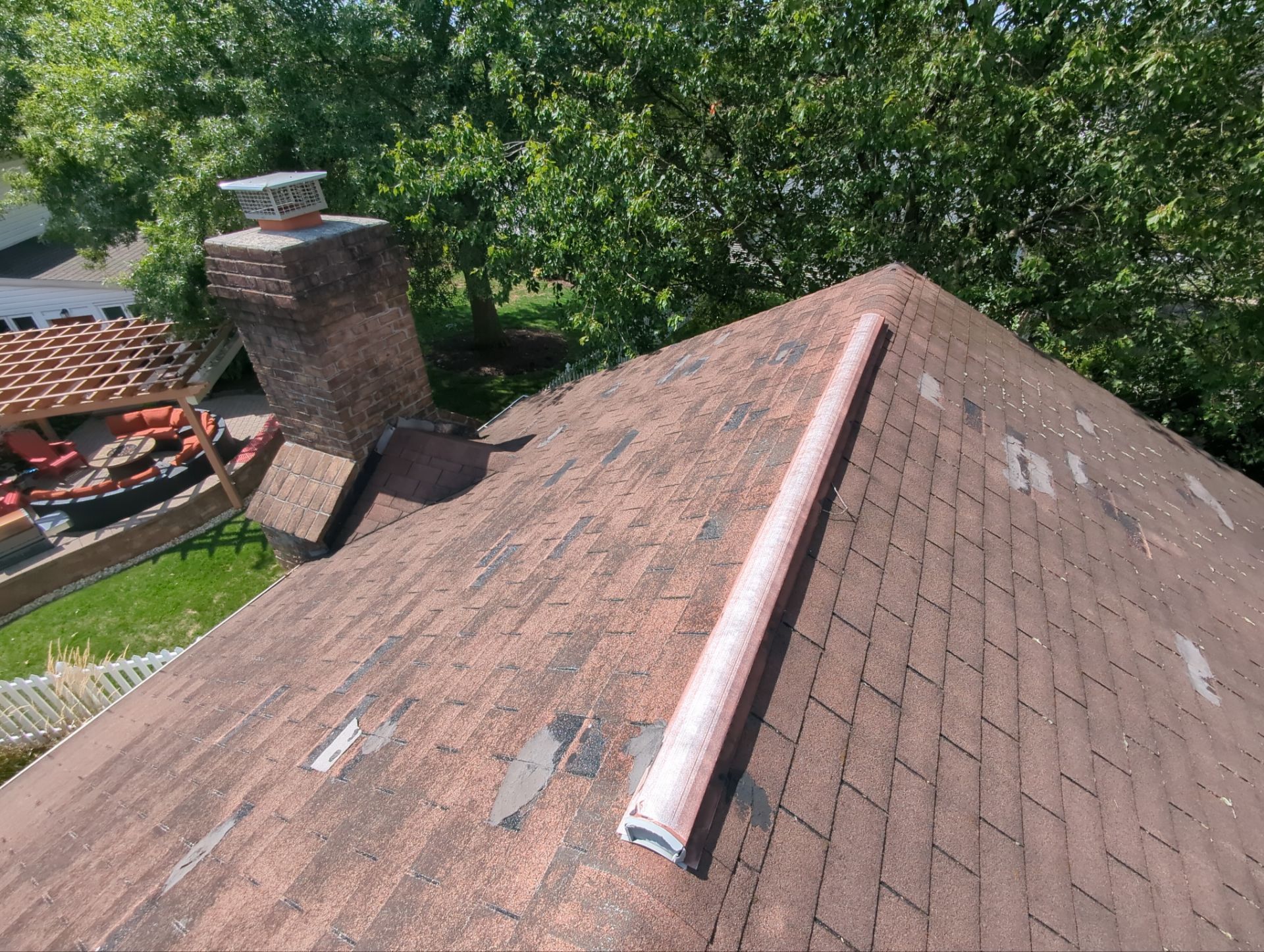 Brown asphalt shingle roof with visible wear, chimney, and metal flashing against a backdrop of green trees.