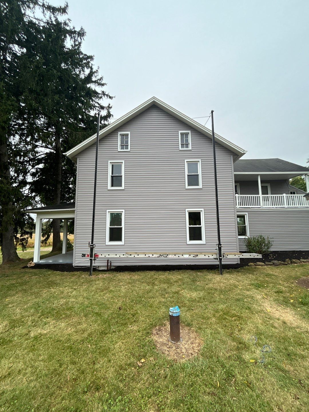 Gray two-story house with siding, six windows, and two poles on a grassy lawn under a cloudy sky.