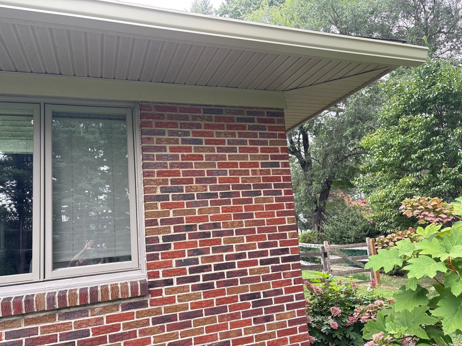 Brick house exterior with a window and overhanging eaves, surrounded by trees and greenery.