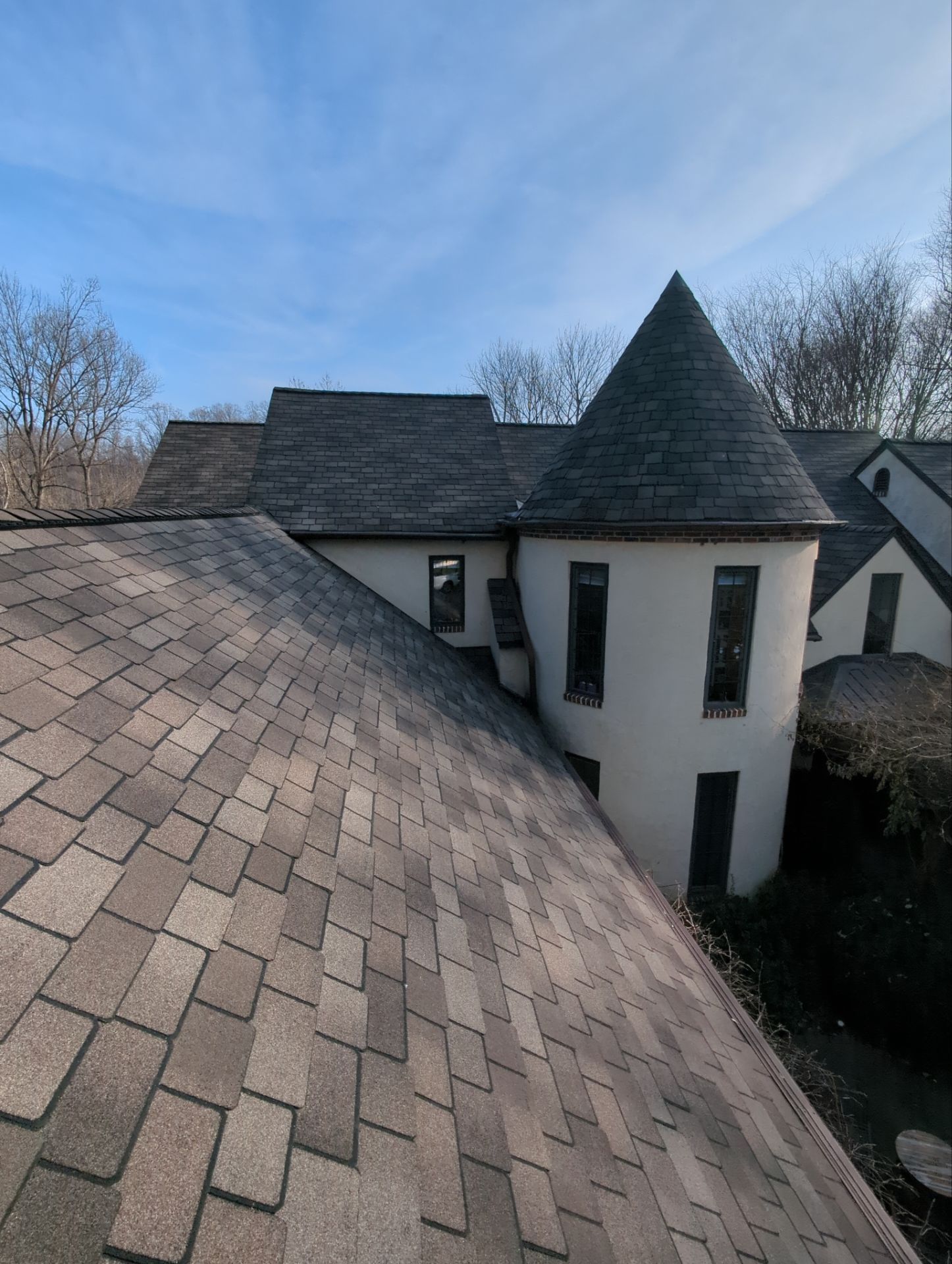 Rooftop view of a light-colored building with dark roof shingles and a conical tower against a blue sky.