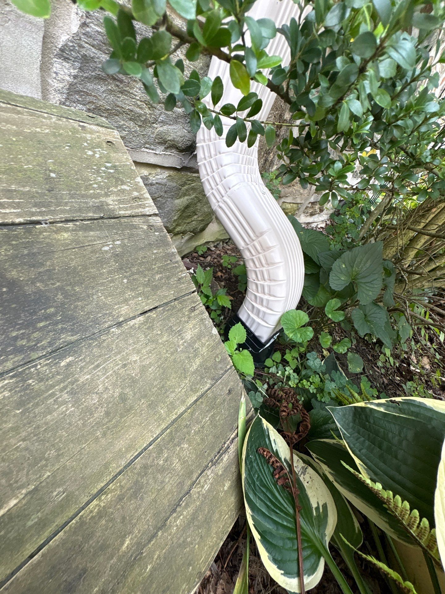 White downspout curves beside a wooden deck, stone wall, and greenery.