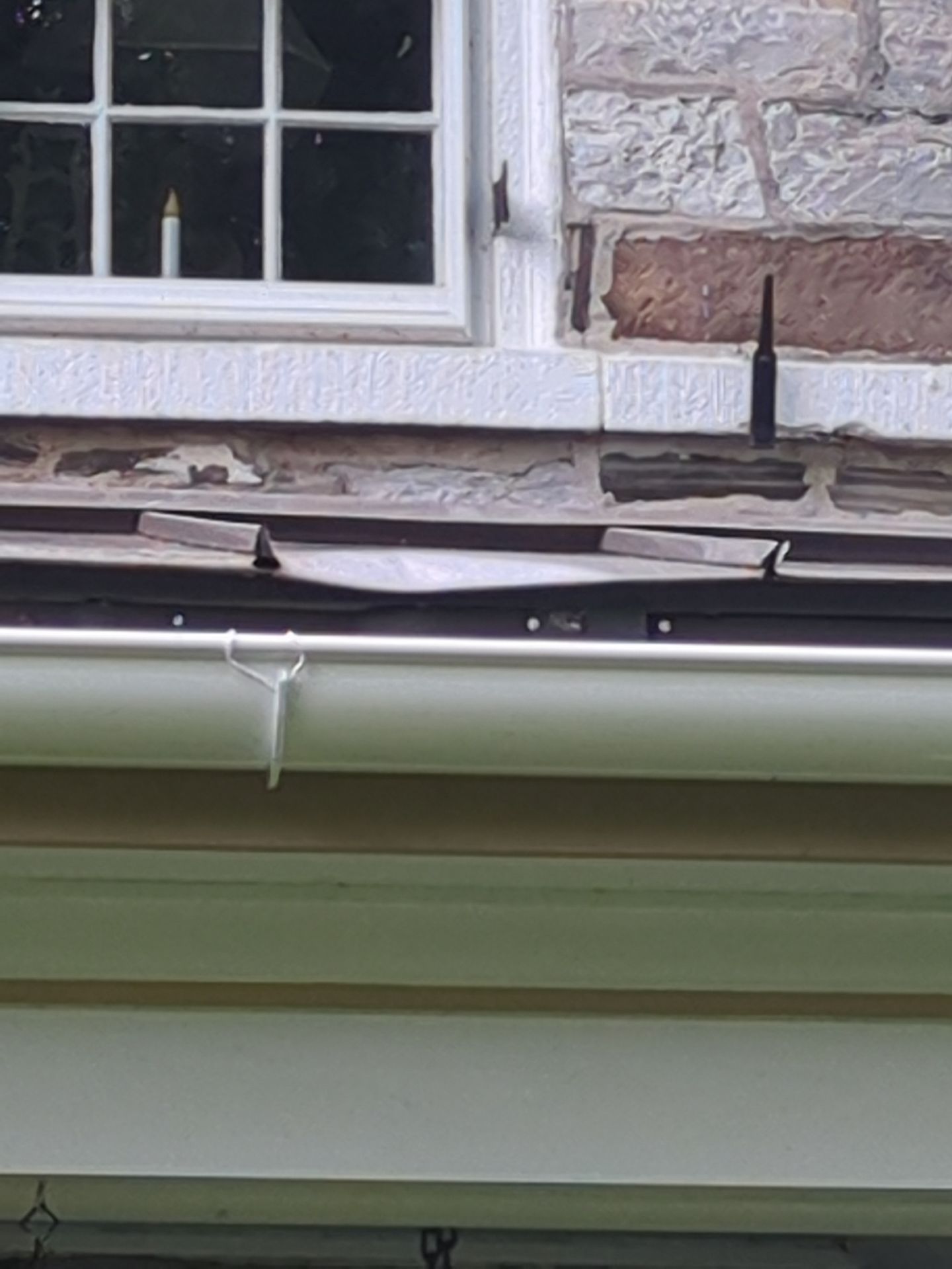 Close-up view of a building's exterior showing a window, stone wall, and a dented metal flashing above a gutter.