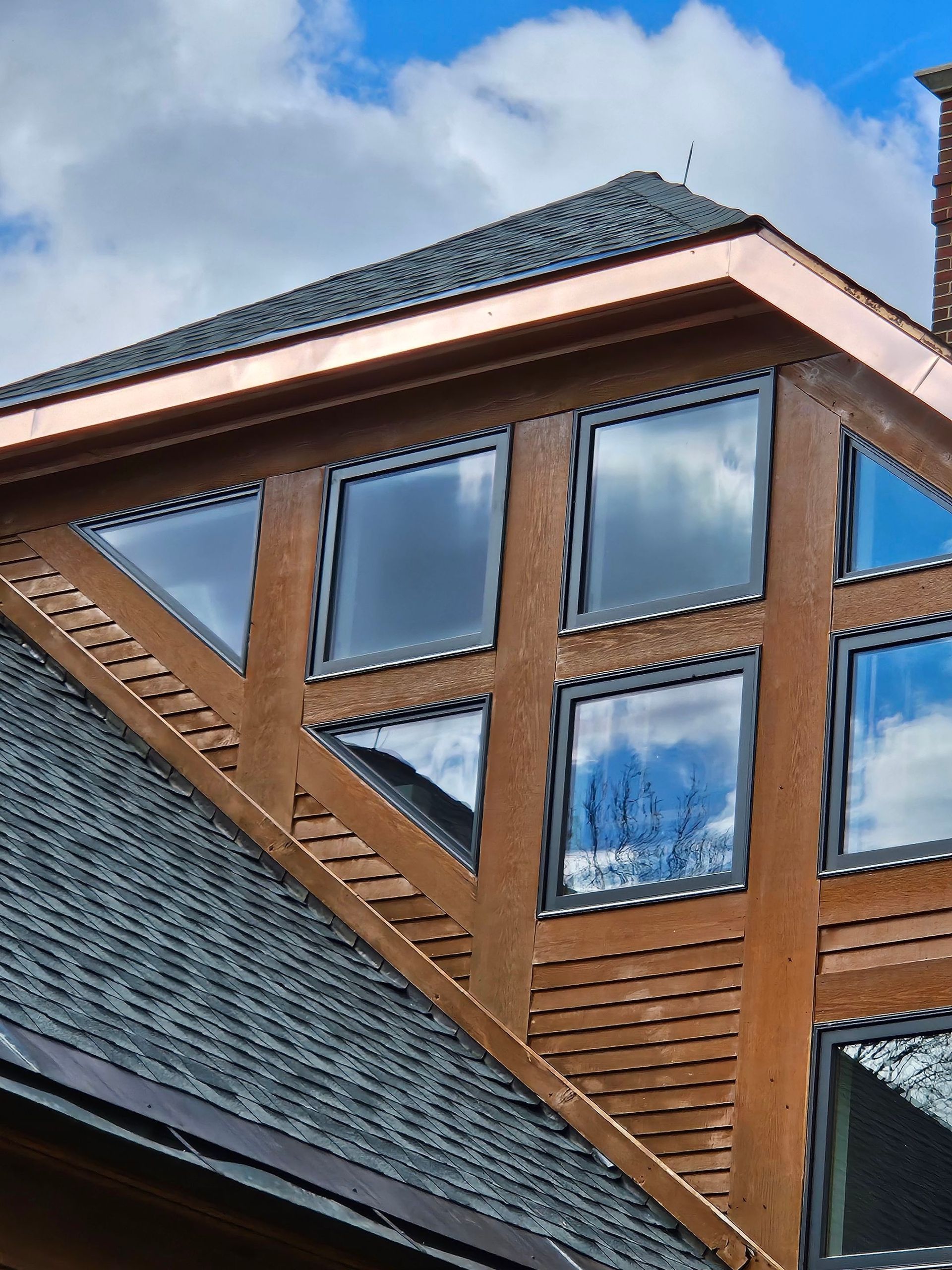 Triangular window structure on a brown wooden exterior with copper trim, under a dark roof, against a blue sky.