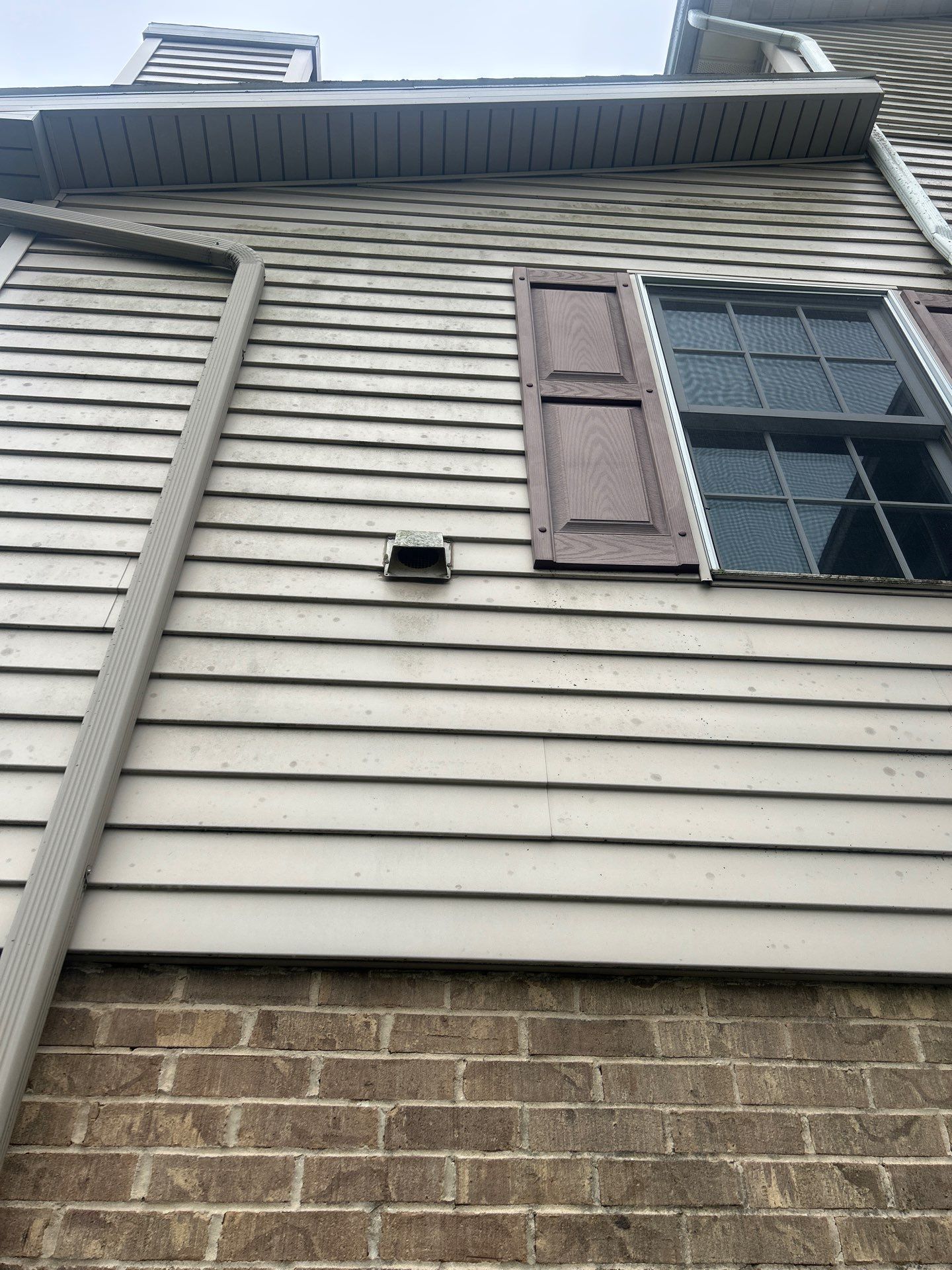 Exterior of a house with beige siding, brick base, window with brown shutters, and vent.