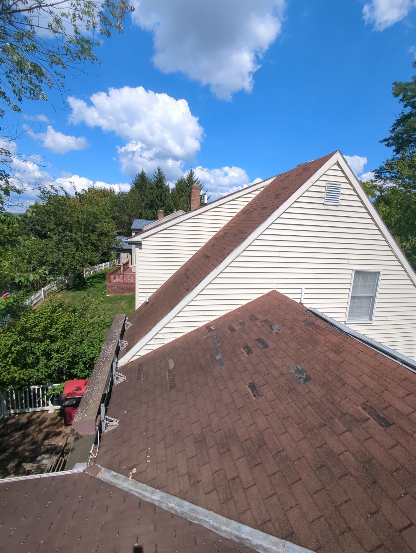 Brown shingled roof of a house with white siding under a bright blue sky with puffy clouds.