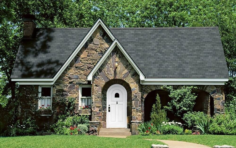 Two-story house with red siding, wrap-around porch, and turret under a gray roof with a flowering tree in front.