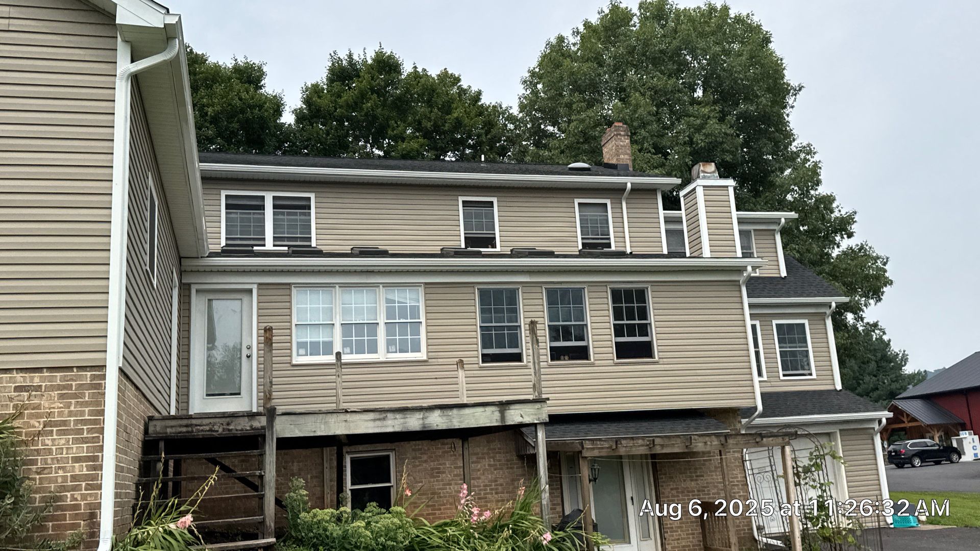 Tan two-story house with a wooden deck, multiple windows, and a brick chimney against a green tree backdrop.