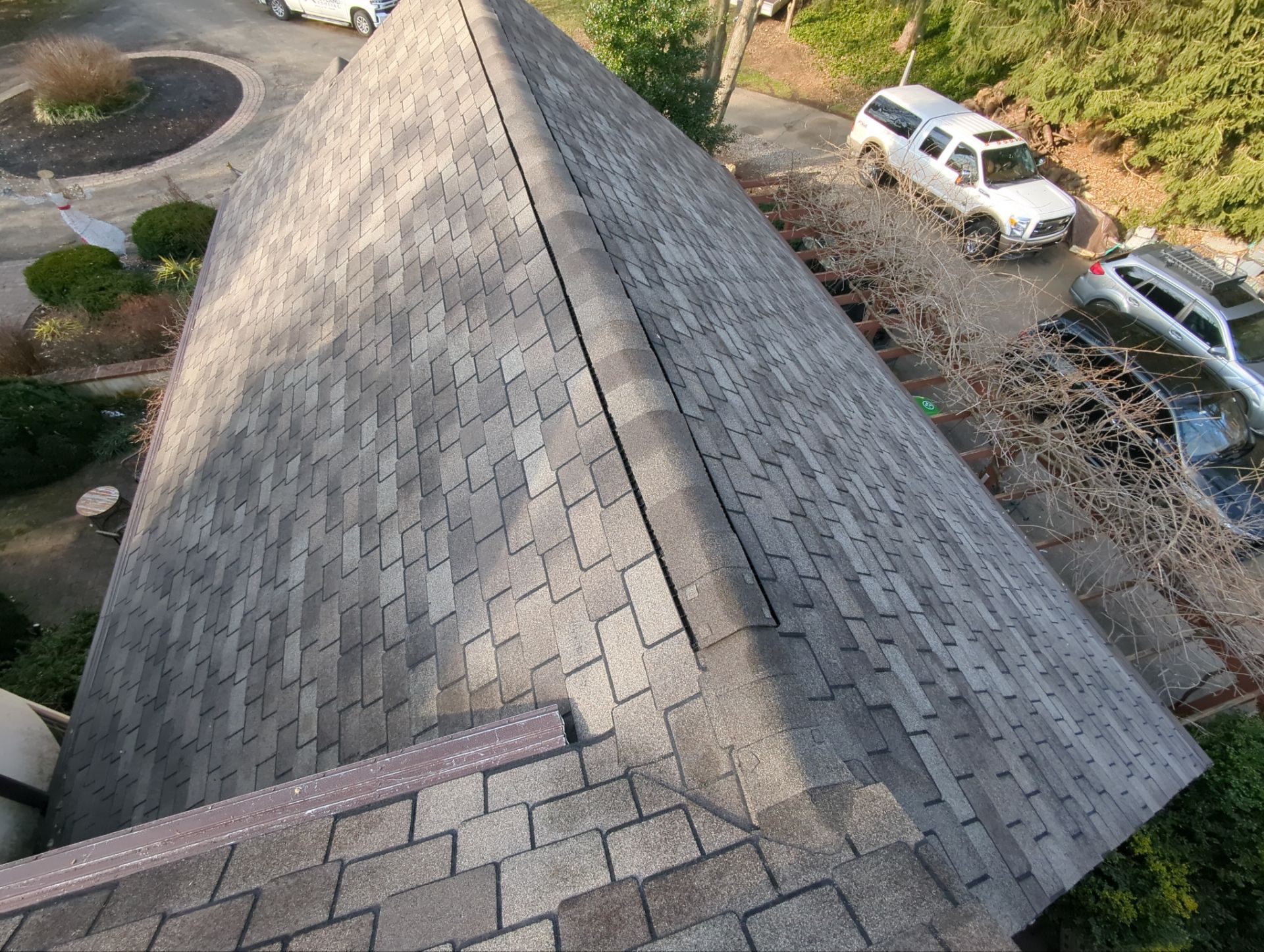 View of a gray shingled roof with a dark ridge line; cars and landscaping visible in the background.