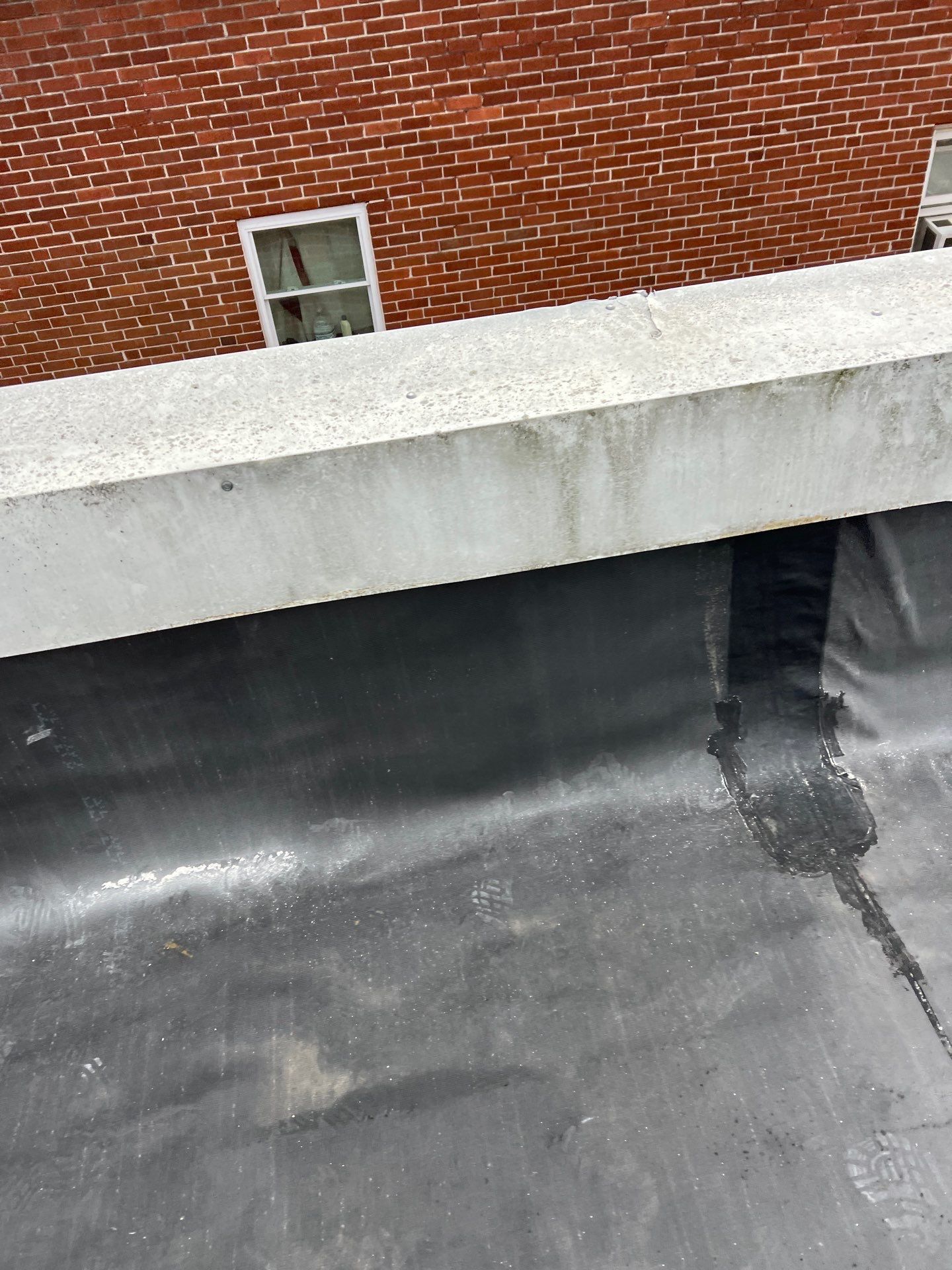 Black roofing material meets a concrete ledge, with a brick building in the background.