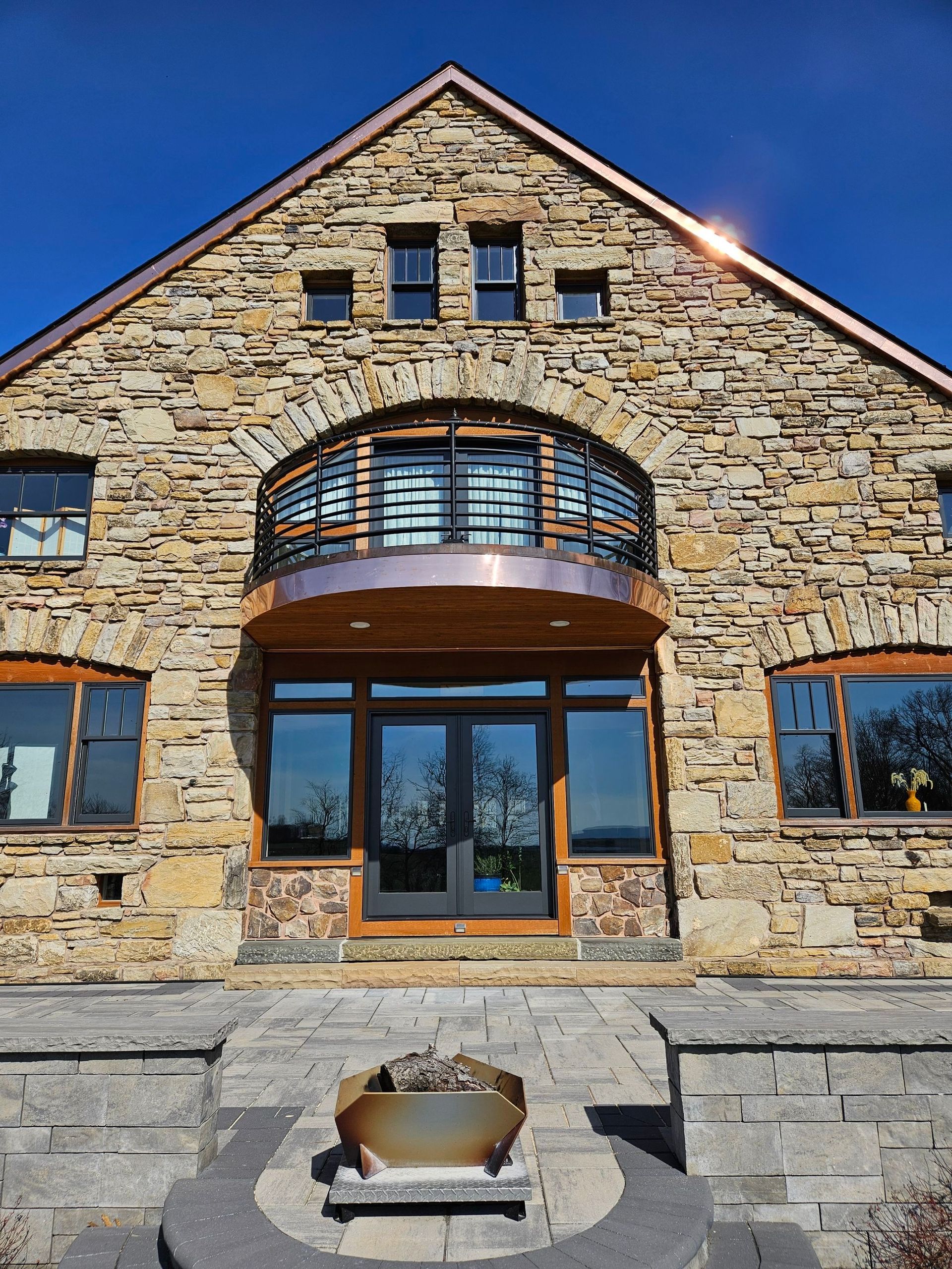 Stone house with arched balcony, copper accents, and large windows against a blue sky.