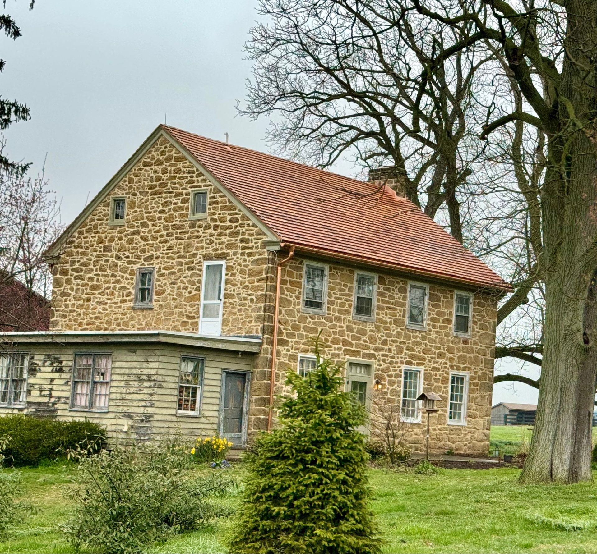 Stone farmhouse with red-tiled roof and weathered porch; green lawn and trees.
