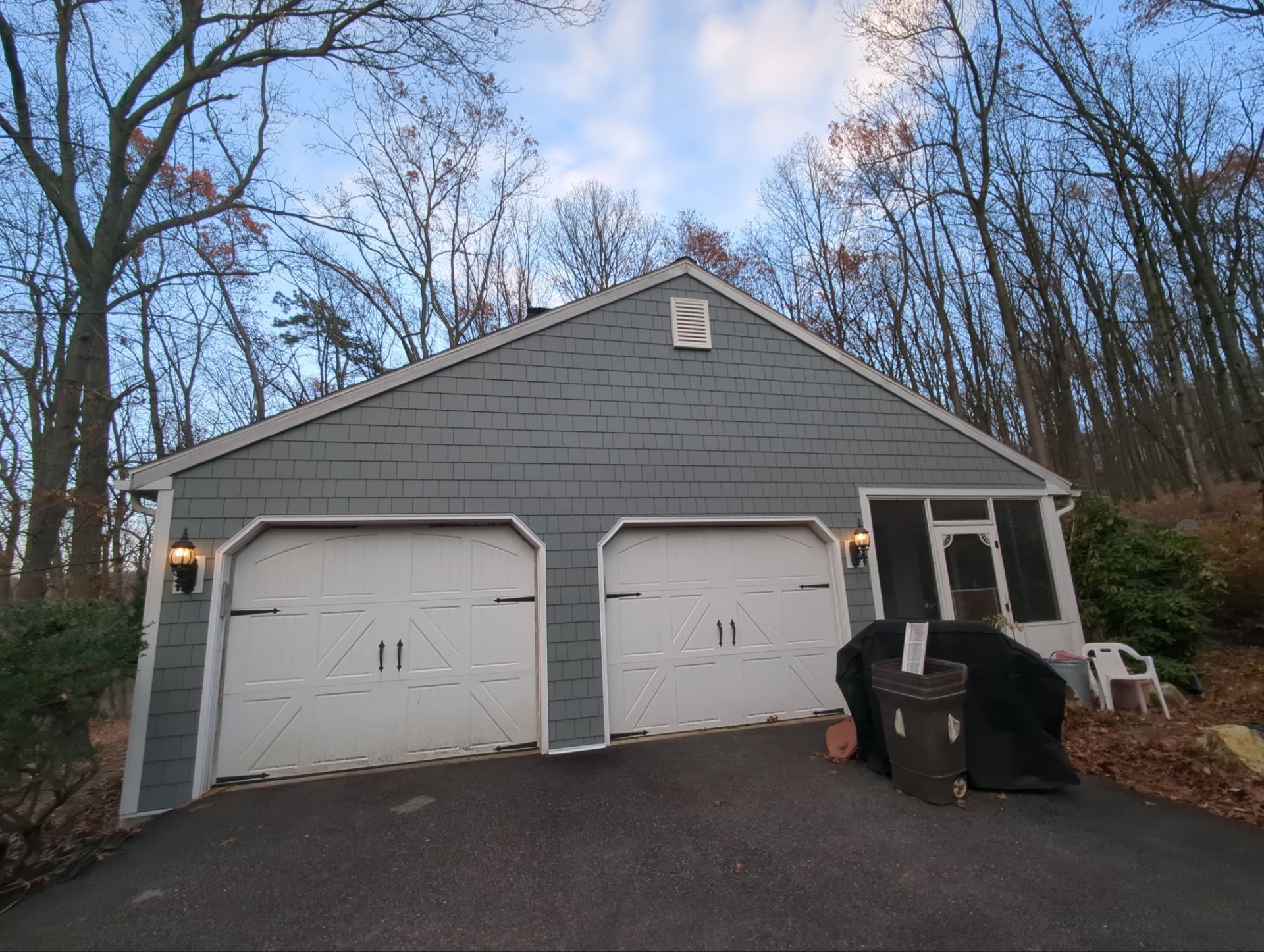 Two-car garage with gray siding and white doors, set on a slightly sloped driveway with trees in the background.