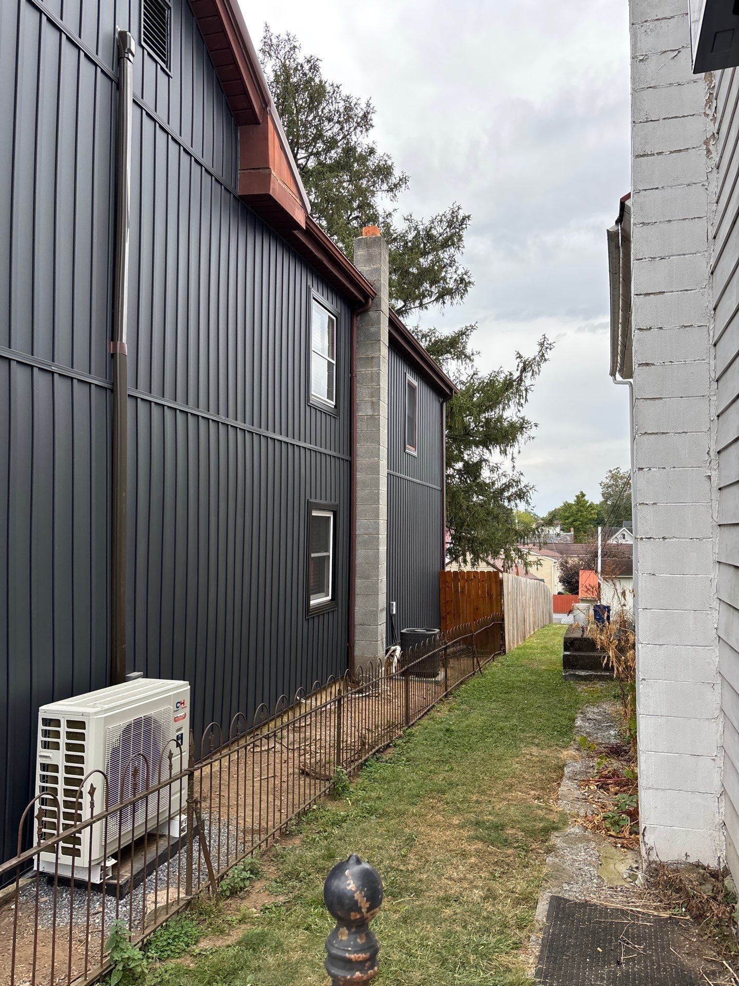 Black-sided building with a chimney, narrow alley with fence, and white building in a cloudy setting.